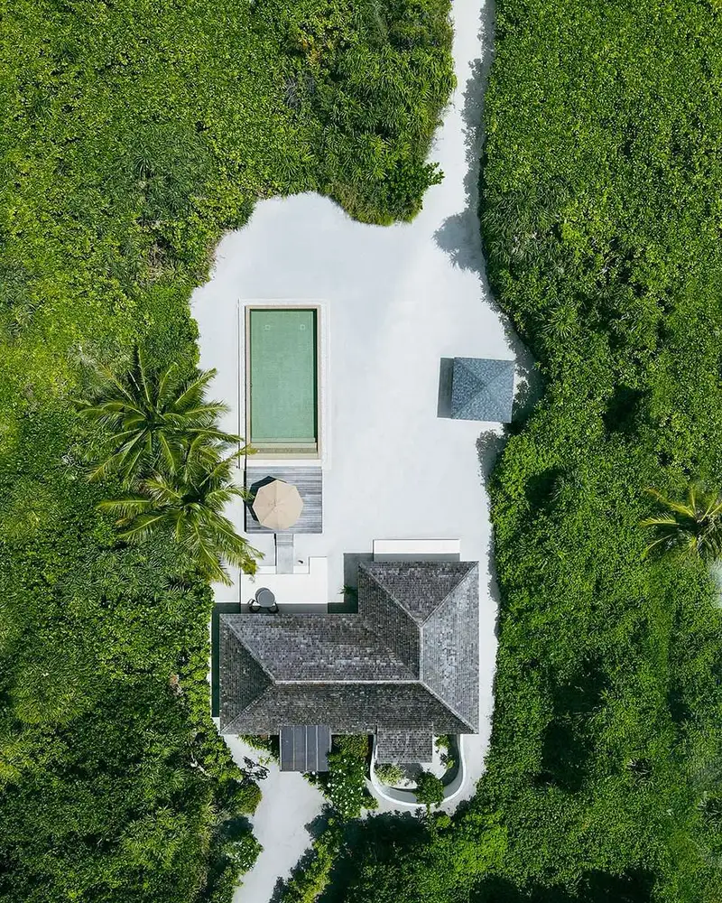 Vertical aerial single thatched villa with long rectangular pool and sandy courtyard.
