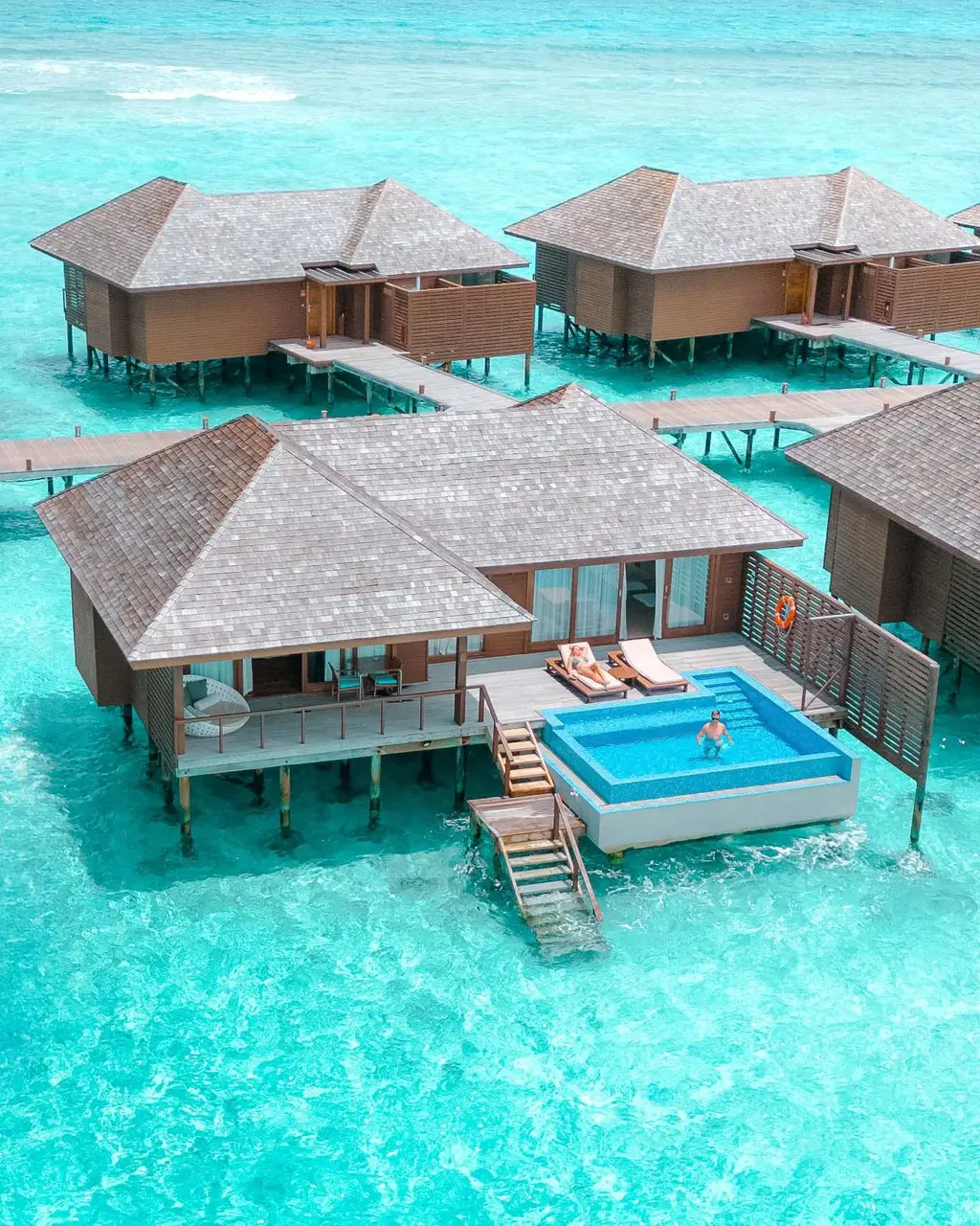 Water villa exterior with deck, rectangular pool, loungers and steps into lagoon; person in pool.