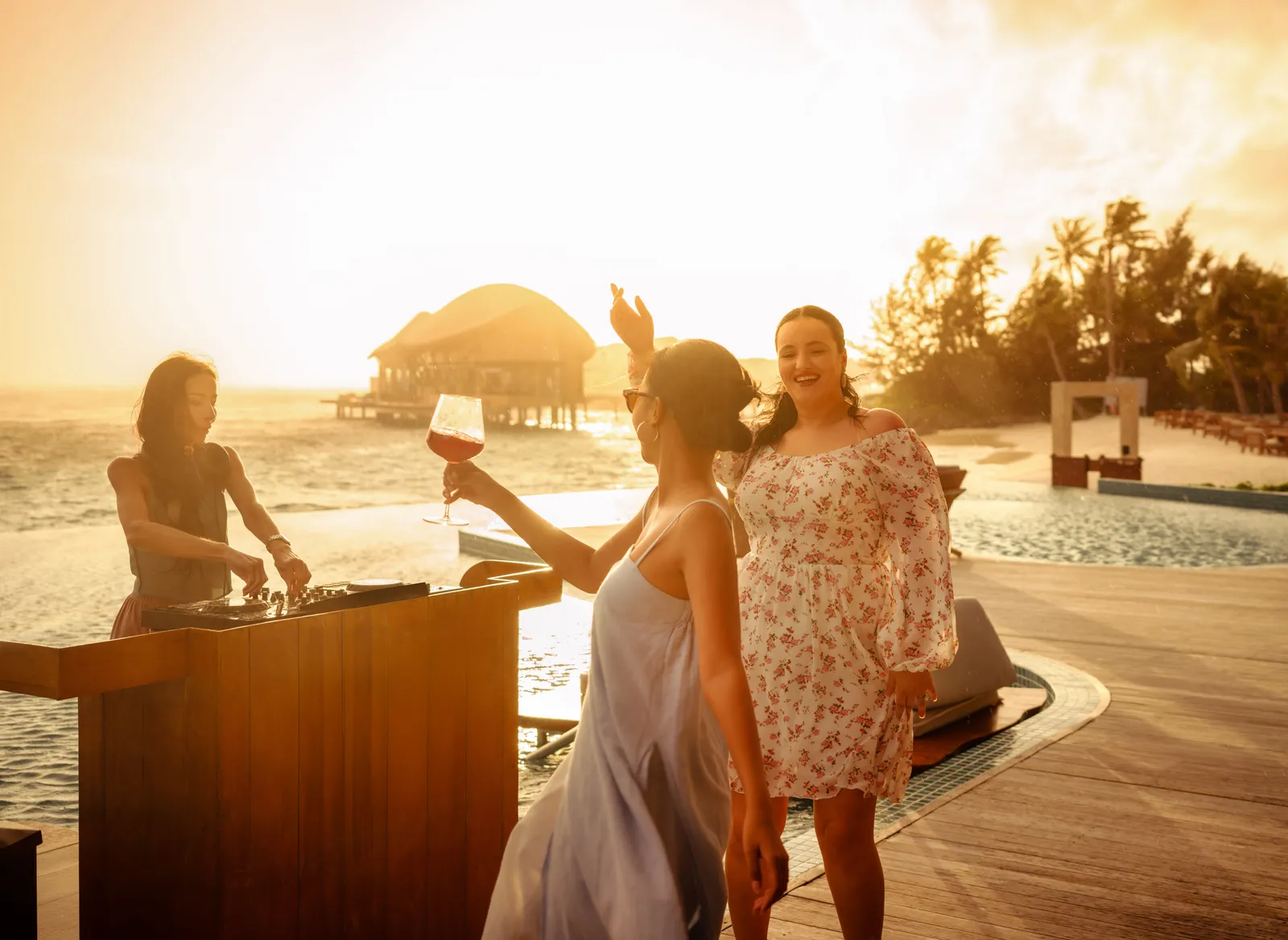 2 friends raise a glass at Aura Pool Bar at sunset, the DJ at the decks and the beach behind.