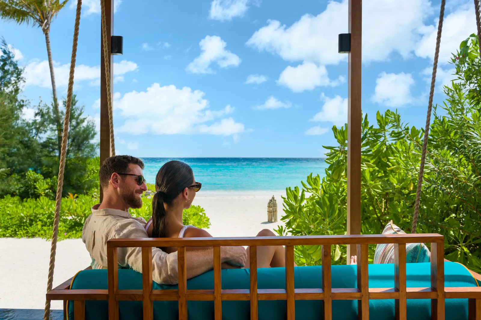 A couple on the swing of a beach villa, framed by greenery, with the sea ahead.