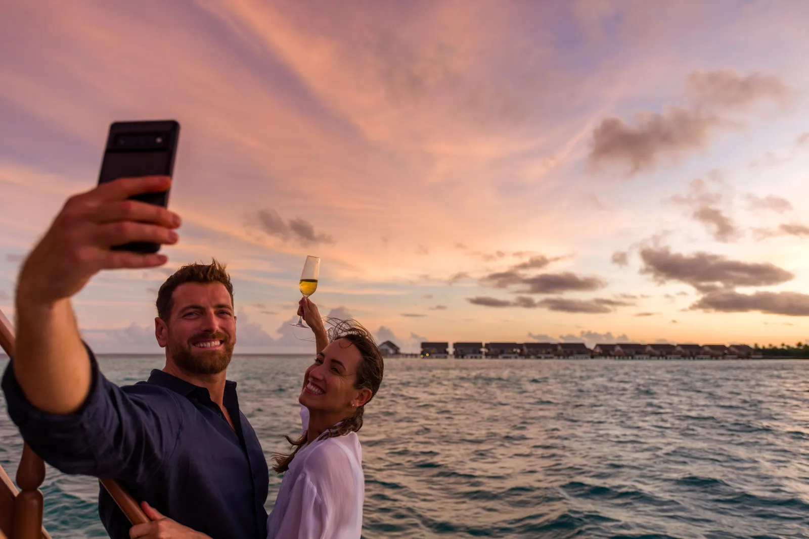 A couple take a photograph of themselves on a dhoni at sunset, the water villas far off.