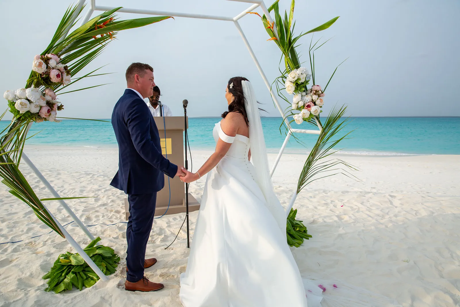 2 sposi si tengono per mano sotto un arco di fiori sulla spiaggia, con la laguna turchese dietro.