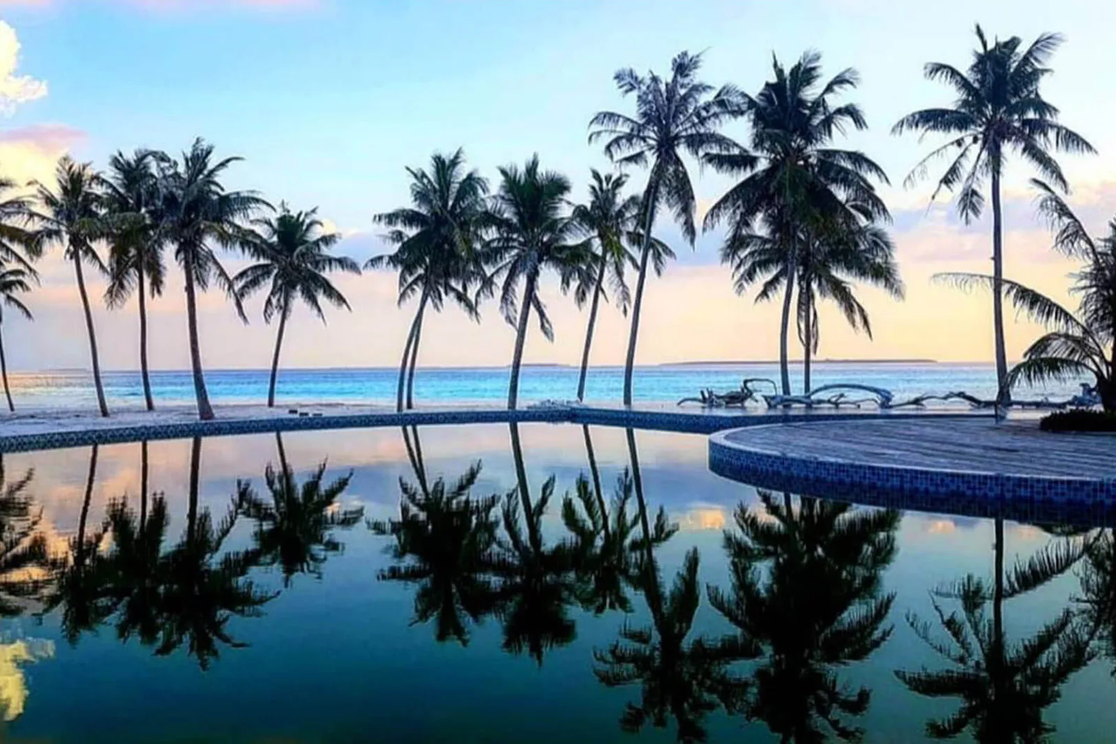 La piscina a sfioro sulla punta dell’isola riflette le palme e il cielo del tramonto, con il mare dietro.