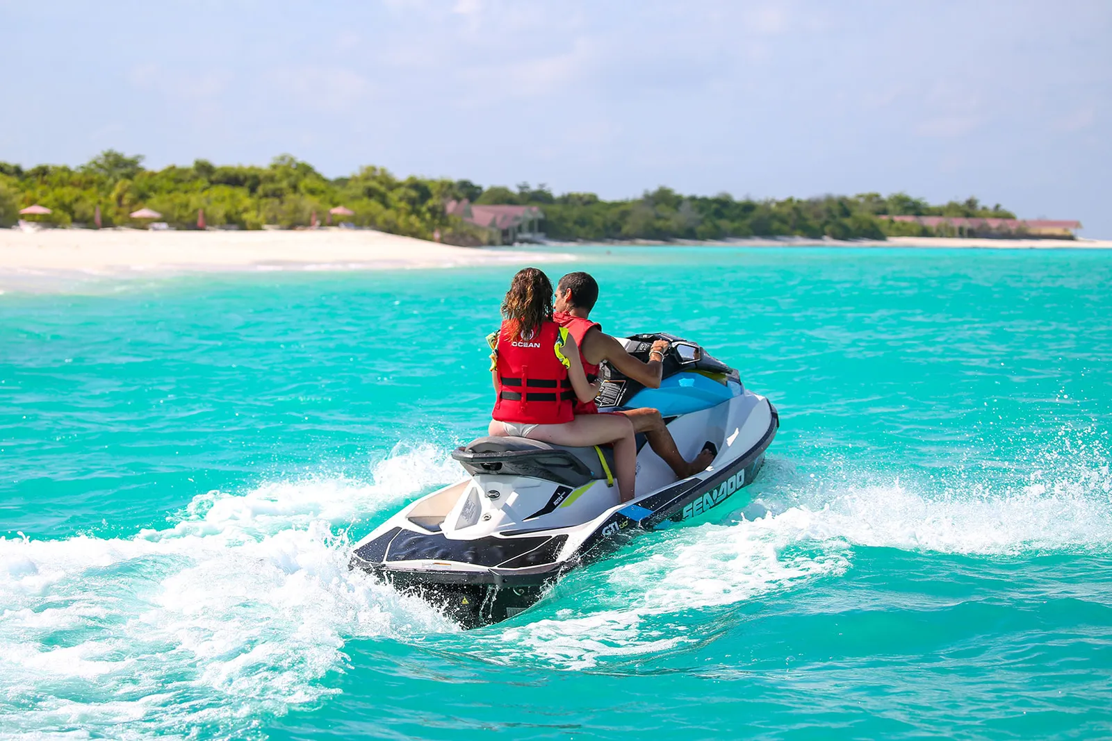 2 persone su una moto d’acqua nella laguna, con l’isola verde e la spiaggia sullo sfondo.