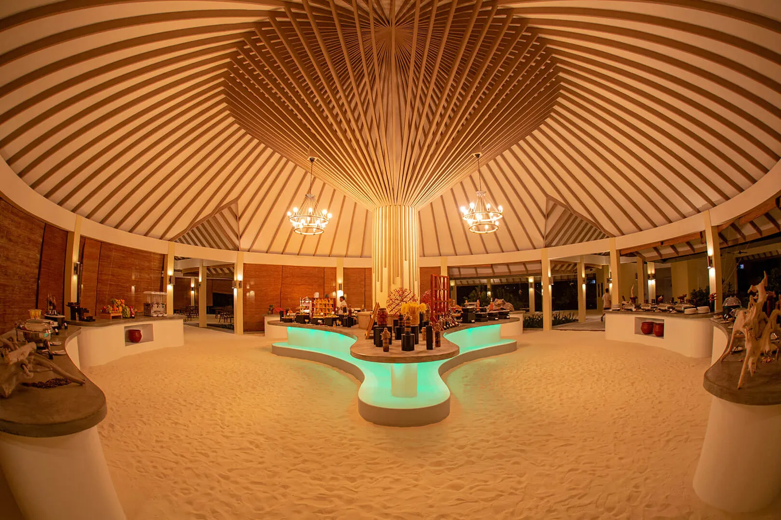 Il Main Restaurant di sera: il grande padiglione rotondo con il soffitto di legno a raggiera, il pavimento di sabbia e il buffet al centro.