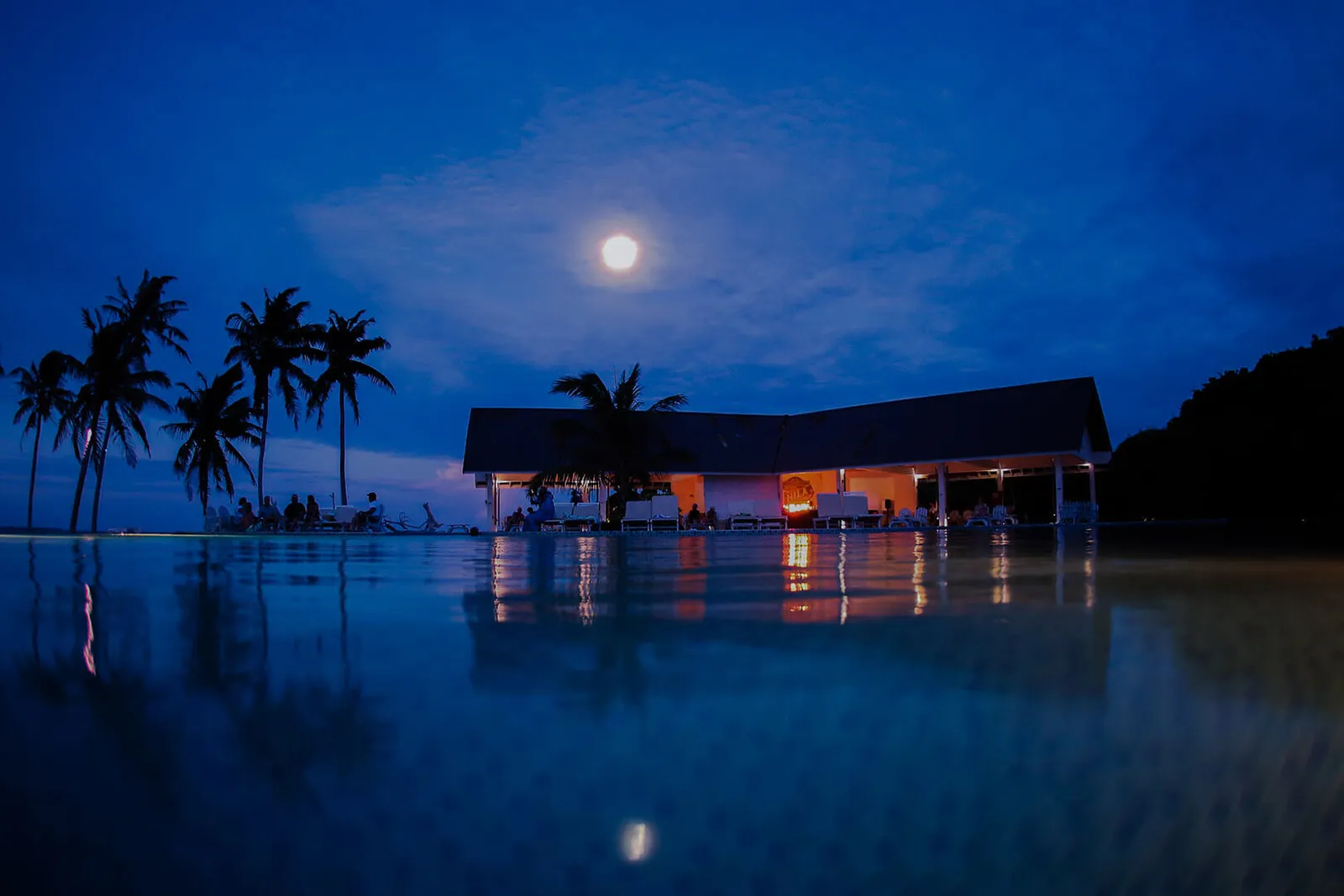 Il padiglione del ristorante illuminato di notte, sotto la luna, riflesso nell’acqua ferma.