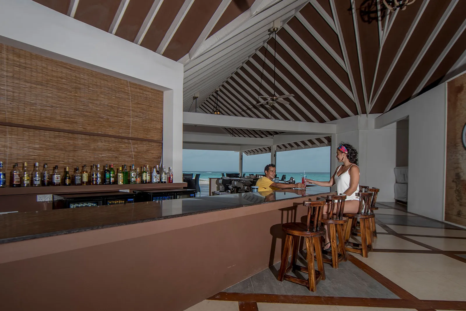 Il banco del Thila Bar, con le bottiglie, gli sgabelli di legno e il mare oltre le colonne.