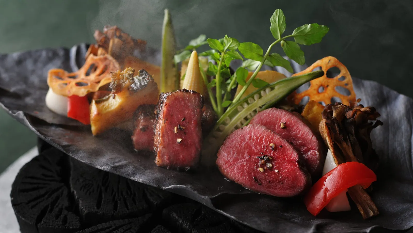 Sliced venison and wagyu with grilled vegetables on a dark plate