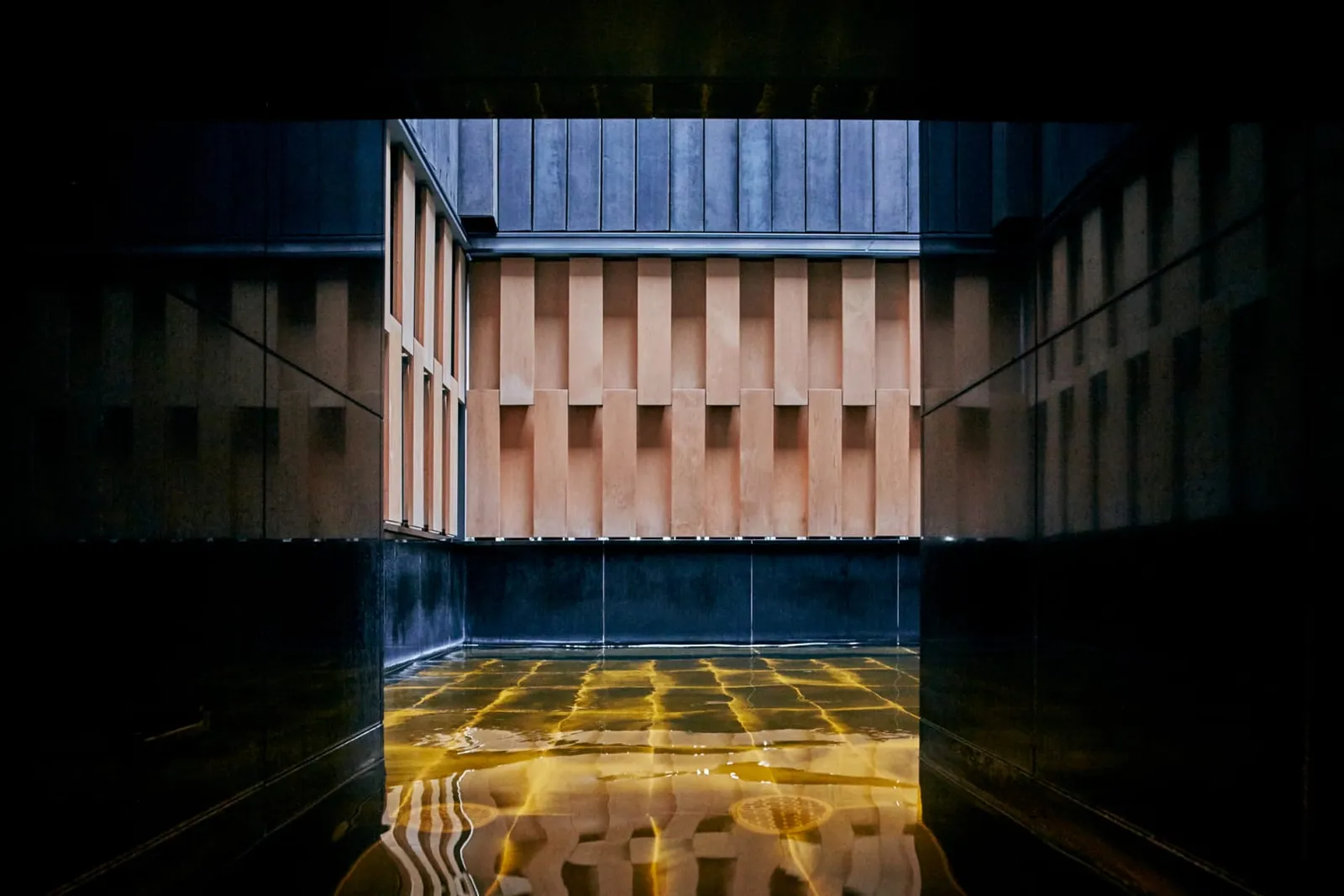 The open-air onsen bath on the top floor: amber water lit from below between timber walls, the sky above.