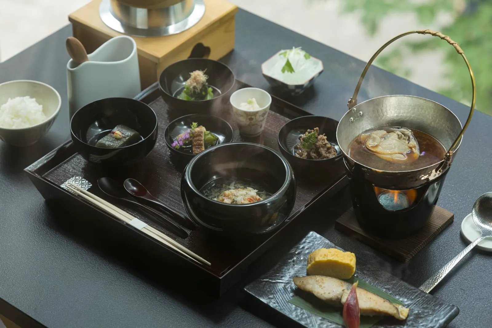 The Japanese breakfast on a black tray: bowls of rice and soup, grilled fish, small dishes and an iron teapot.