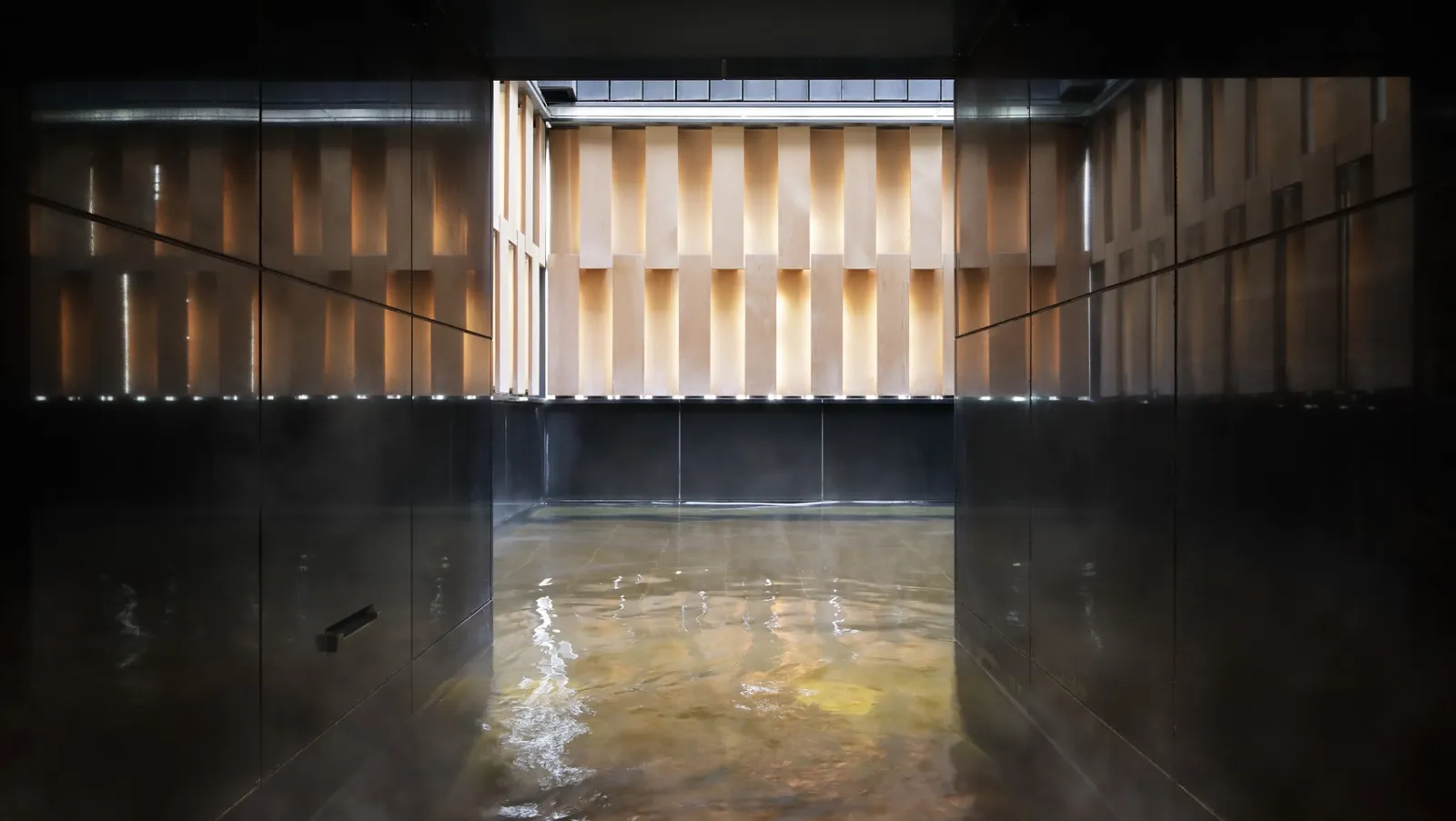 The indoor onsen bath, dark and steaming, opening towards the open-air bath closed by timber slats.