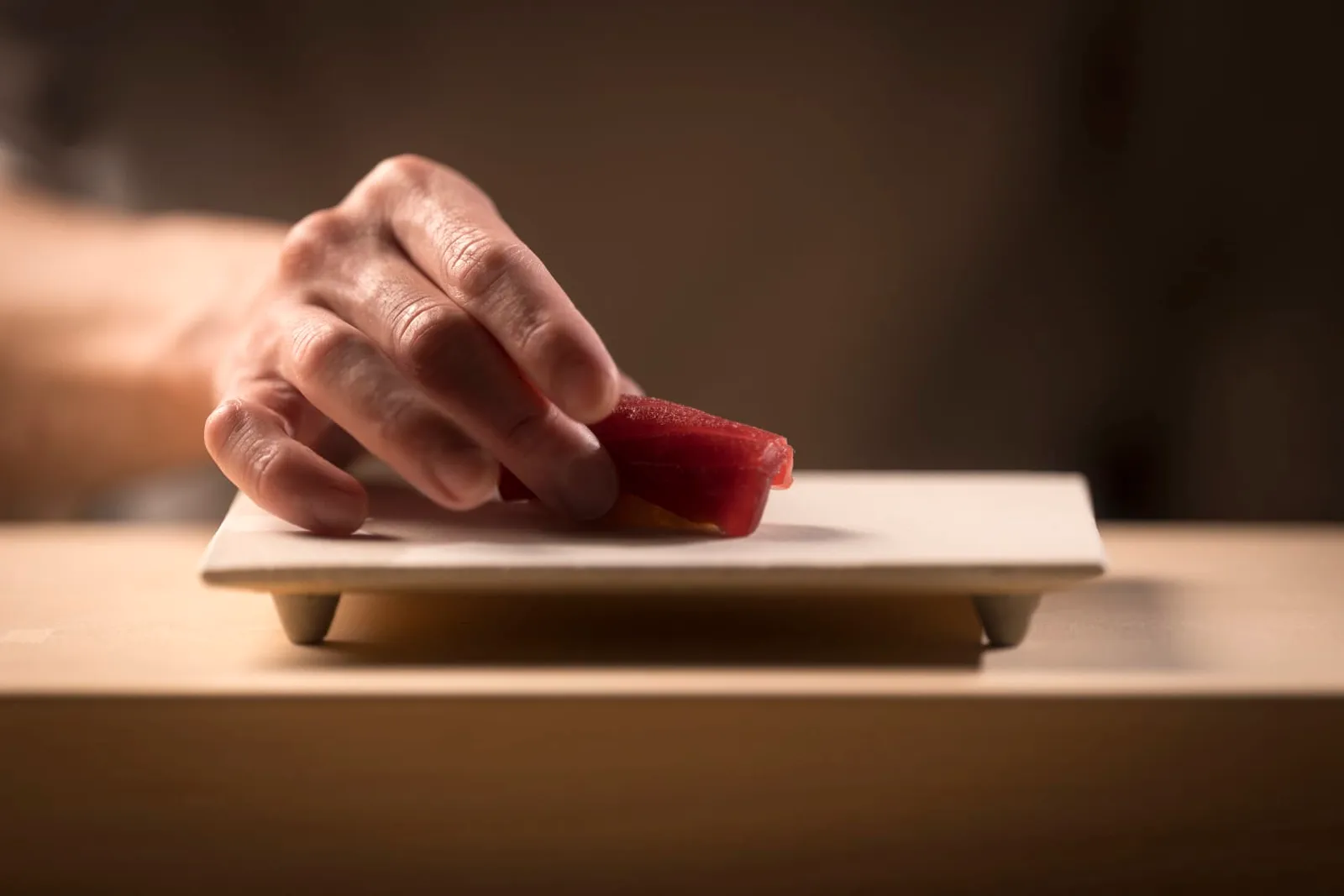 At the Sushi Otemon counter the chef’s hand sets a tuna nigiri on a white plate.