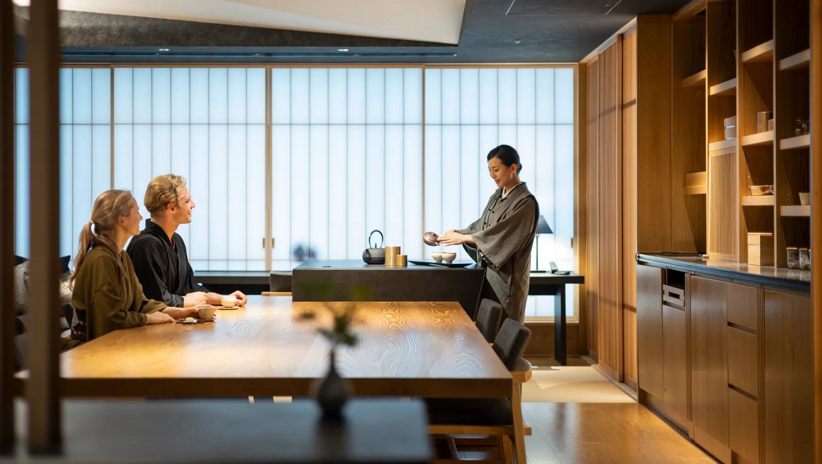 A floor’s OCHANOMA Lounge: 2 guests in kimono at the timber table while a host pours green tea, rice-paper screens behind.