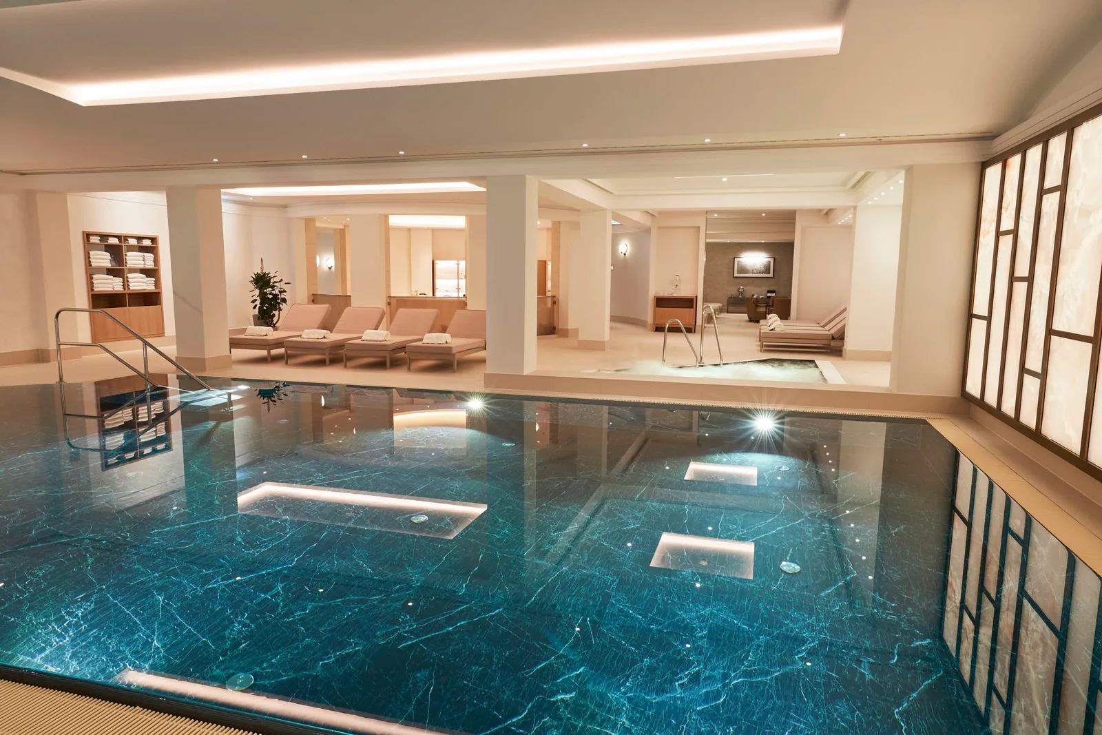 The indoor pool: the basin lined in dark veined stone, lights on the floor, pale loungers along the edge and a wall of glowing panels.