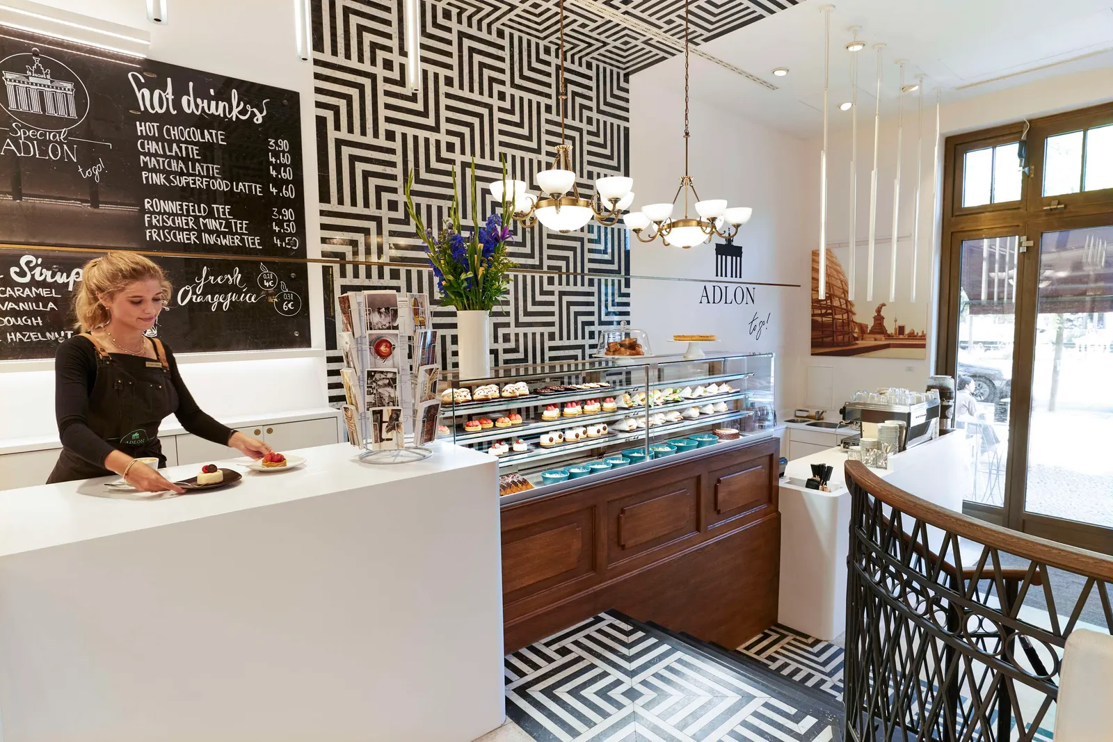 The Adlon To Go coffee shop: a barista serving a pastry at the white counter, the cake display, the drinks blackboard and black-and-white patterned walls.