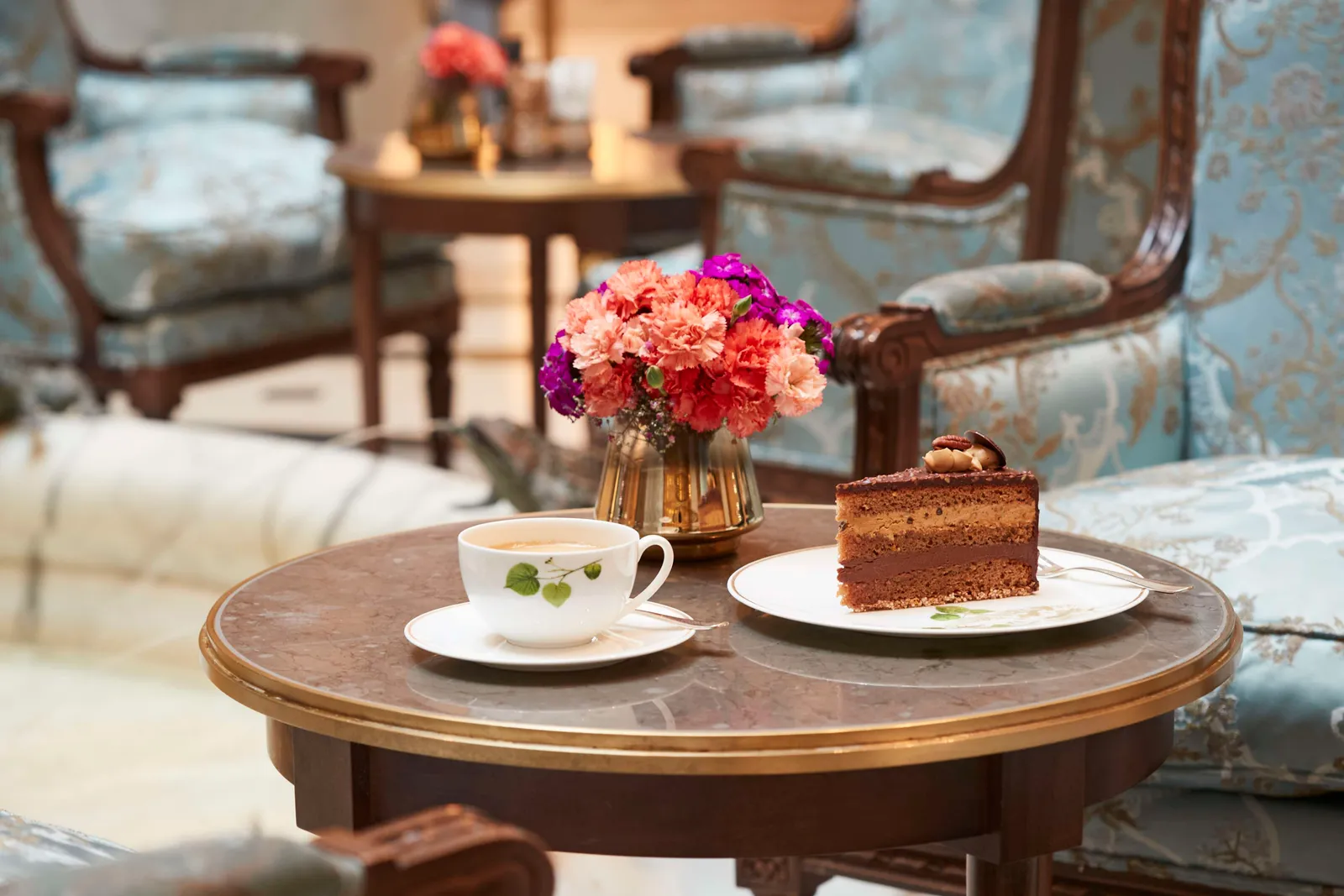 A slice of chocolate Adlon cake and a cup of coffee on a small round lobby table, among blue armchairs and a vase of flowers.