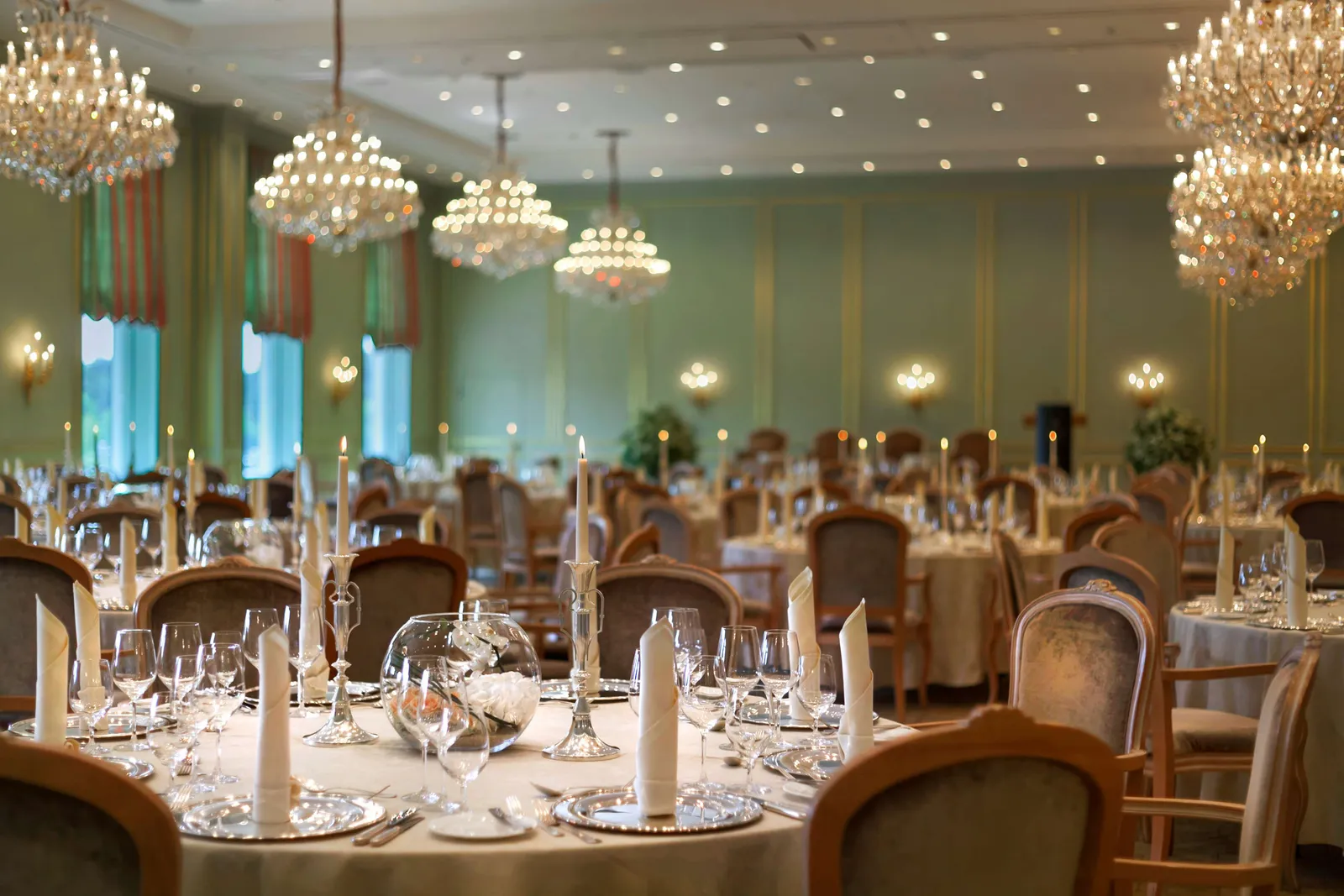 The ballroom laid for a dinner: round tables with candlesticks and glasses, pale-green walls and large crystal chandeliers.