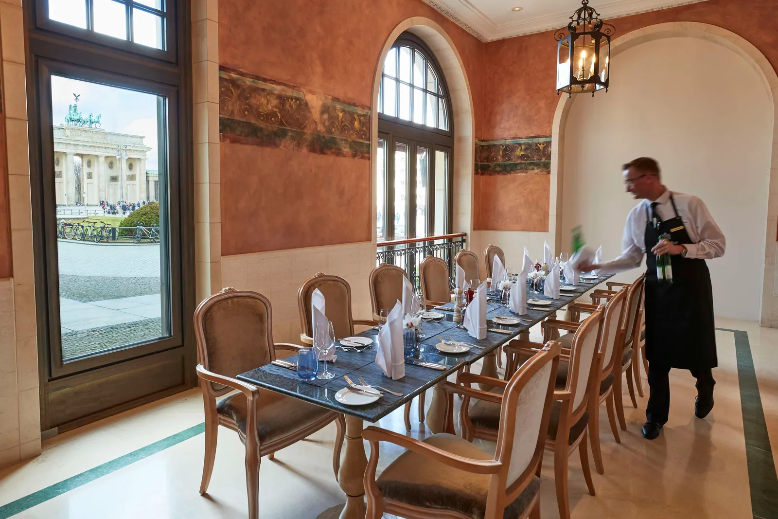 A room of Brasserie Quarré: a long laid table, a waiter at work, terracotta walls and the Brandenburg Gate framed in the window.