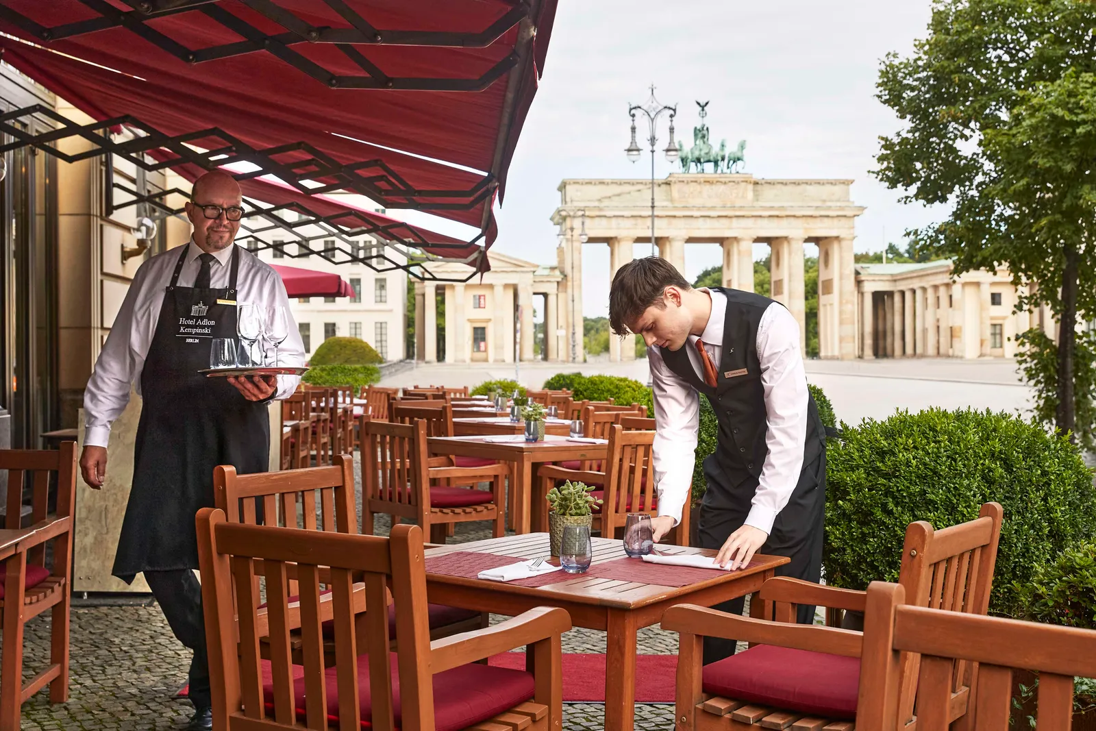 Quarré’s terrace beneath red awnings, with 2 waiters and the Brandenburg Gate beyond the tables.