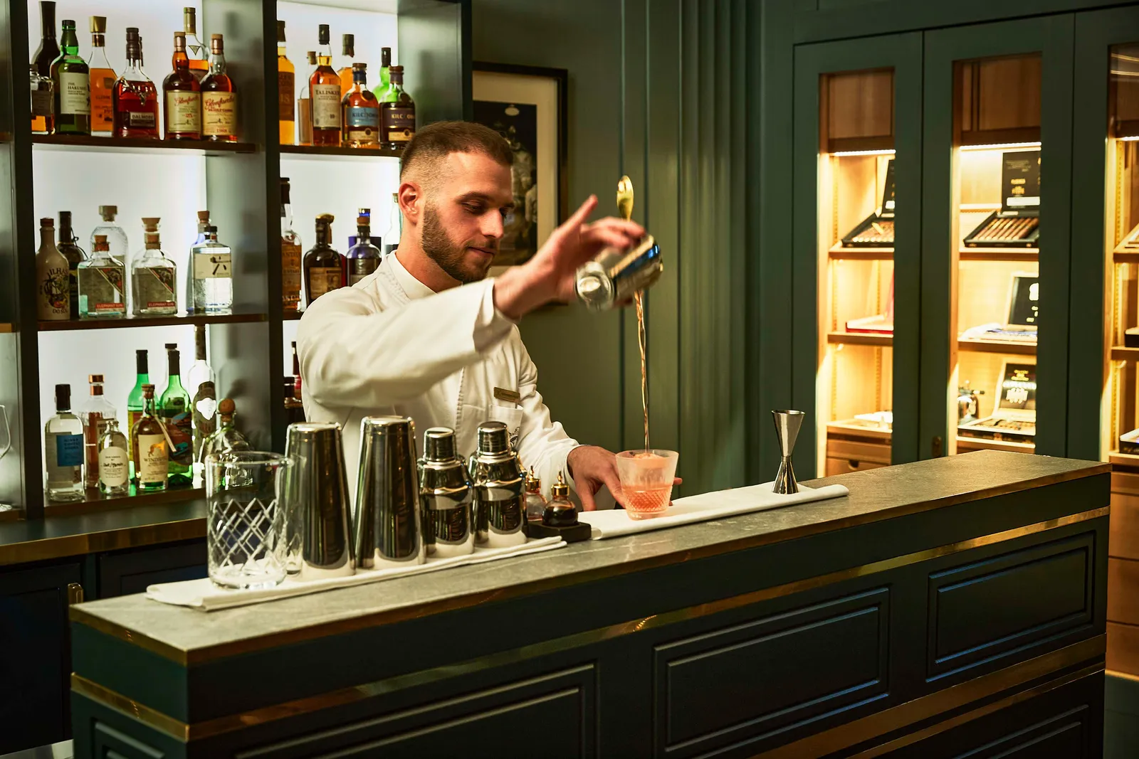 The Elephant Bar counter: a barman in a white jacket pouring a cocktail from the shaker, between shelves of bottles and lit humidors.