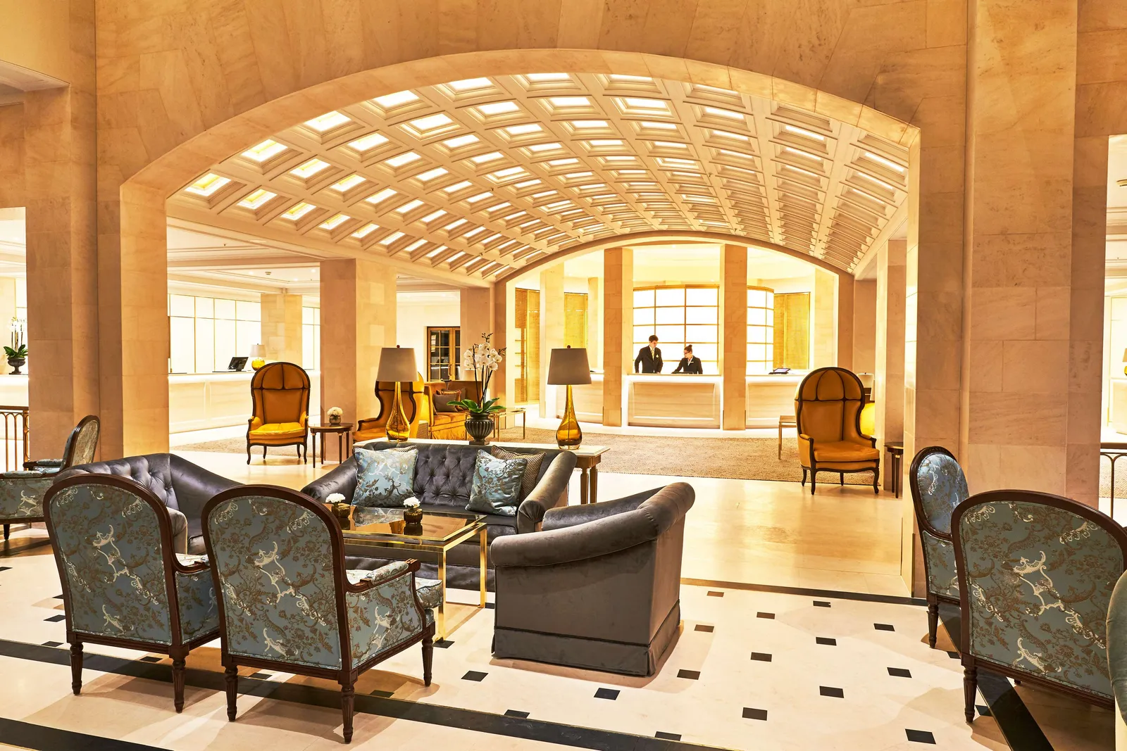 The Lobby Lounge under its coffered vault: grey sofas and blue armchairs on a pale floor with black insets, the reception desk beyond.