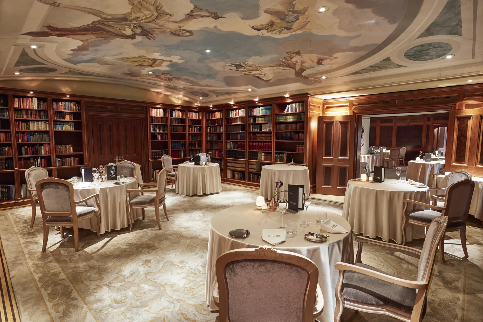 The library room of Lorenz Adlon Esszimmer: round tables with long cloths, wooden bookcases along the walls and a ceiling painted with figures and clouds.