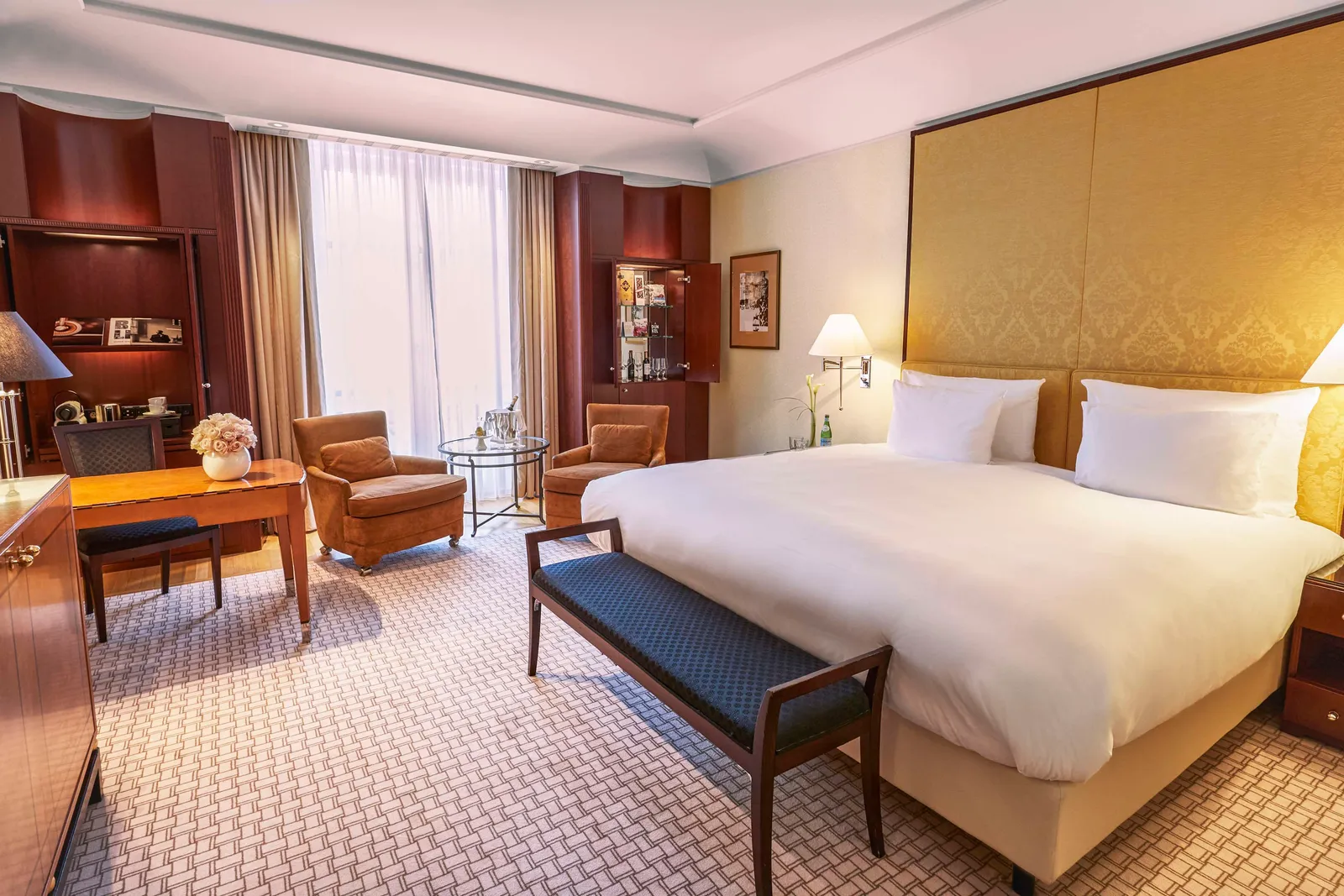 Deluxe Room with a double bed, gold-toned headboard, desk and dark bench.