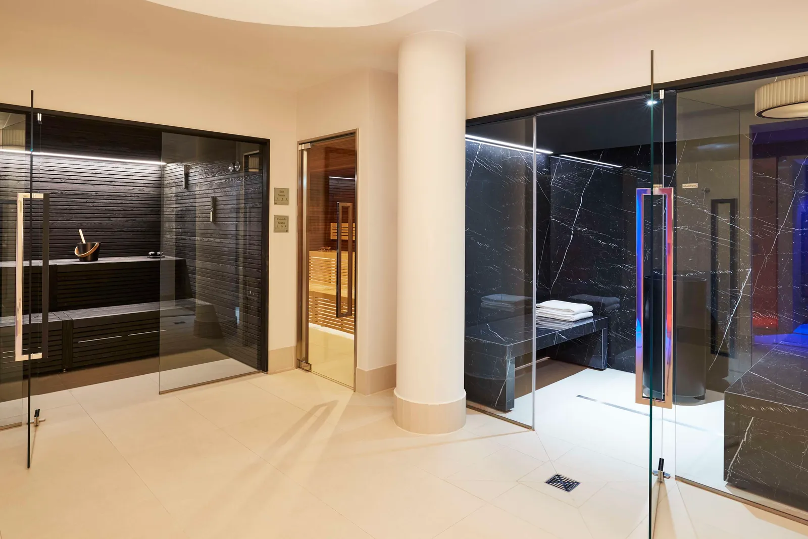 The sauna area: cabins with glass doors, dark stone walls, a white column and a pile of towels on a bench.