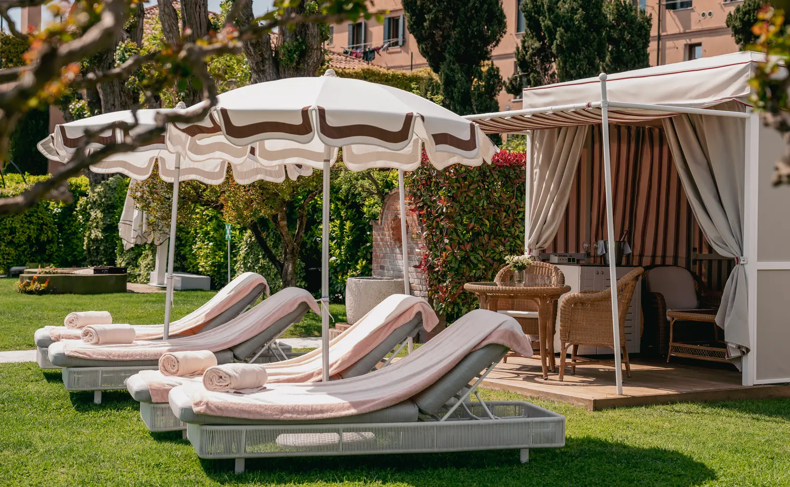 Pink-striped loungers under parasols and a white-curtained cabana on the lawn beside the pool.