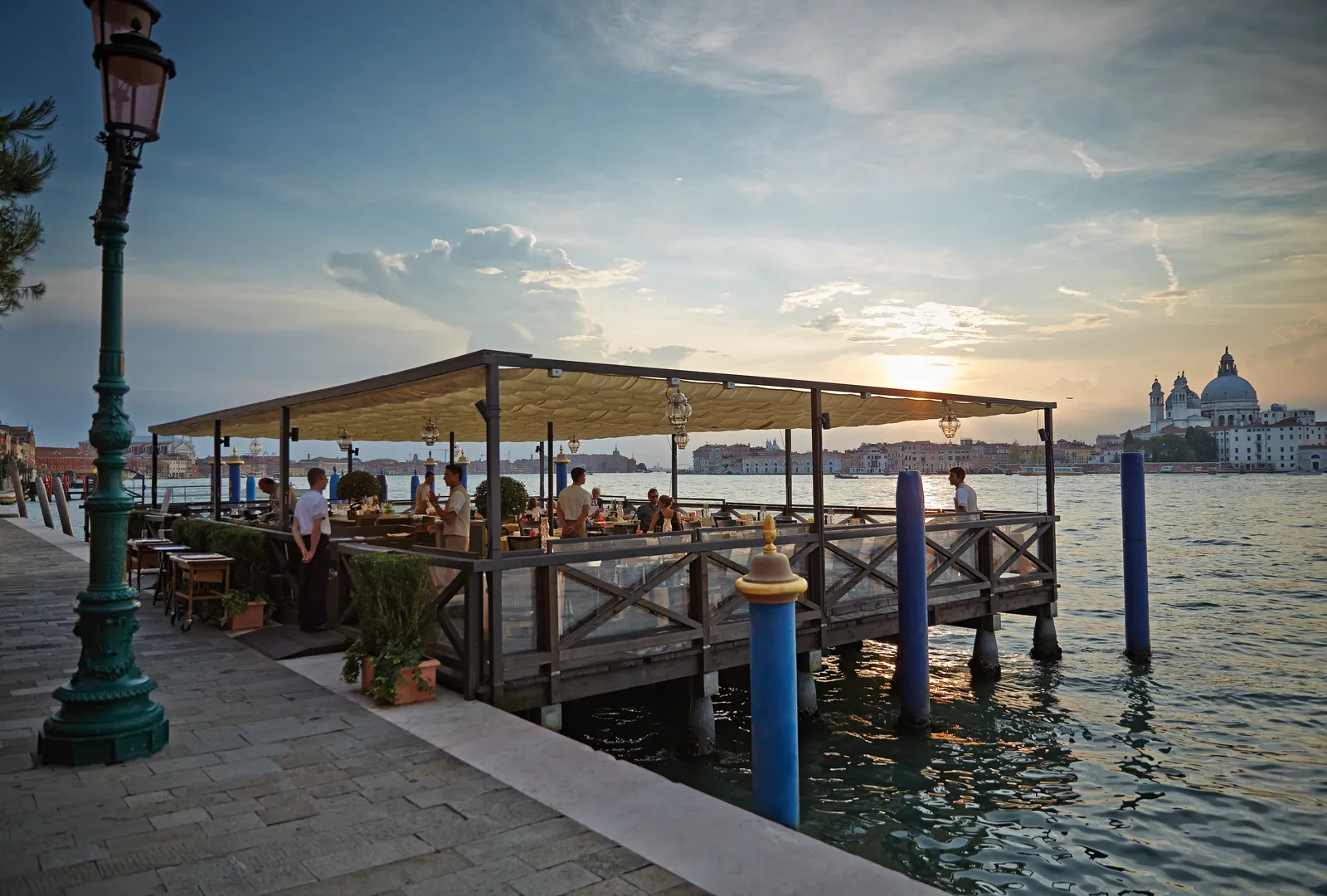 Cip’s Club’s wooden deck over the water at dusk, tables laid and the dome of the Salute in the distance.