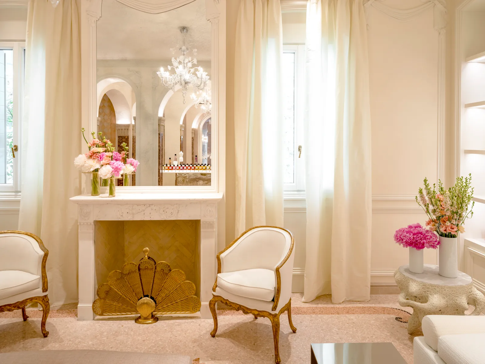 The Dior Spa’s lounge: white armchairs edged in gold, a fireplace with a fan-shaped screen and pale curtains at the windows.