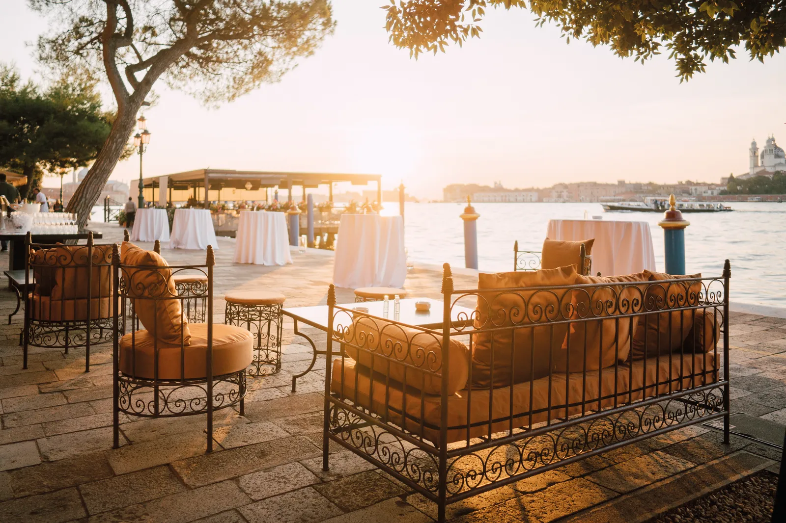 The waterside terrace at sunset, wrought-iron sofas under the pines and the lagoon towards the Salute.