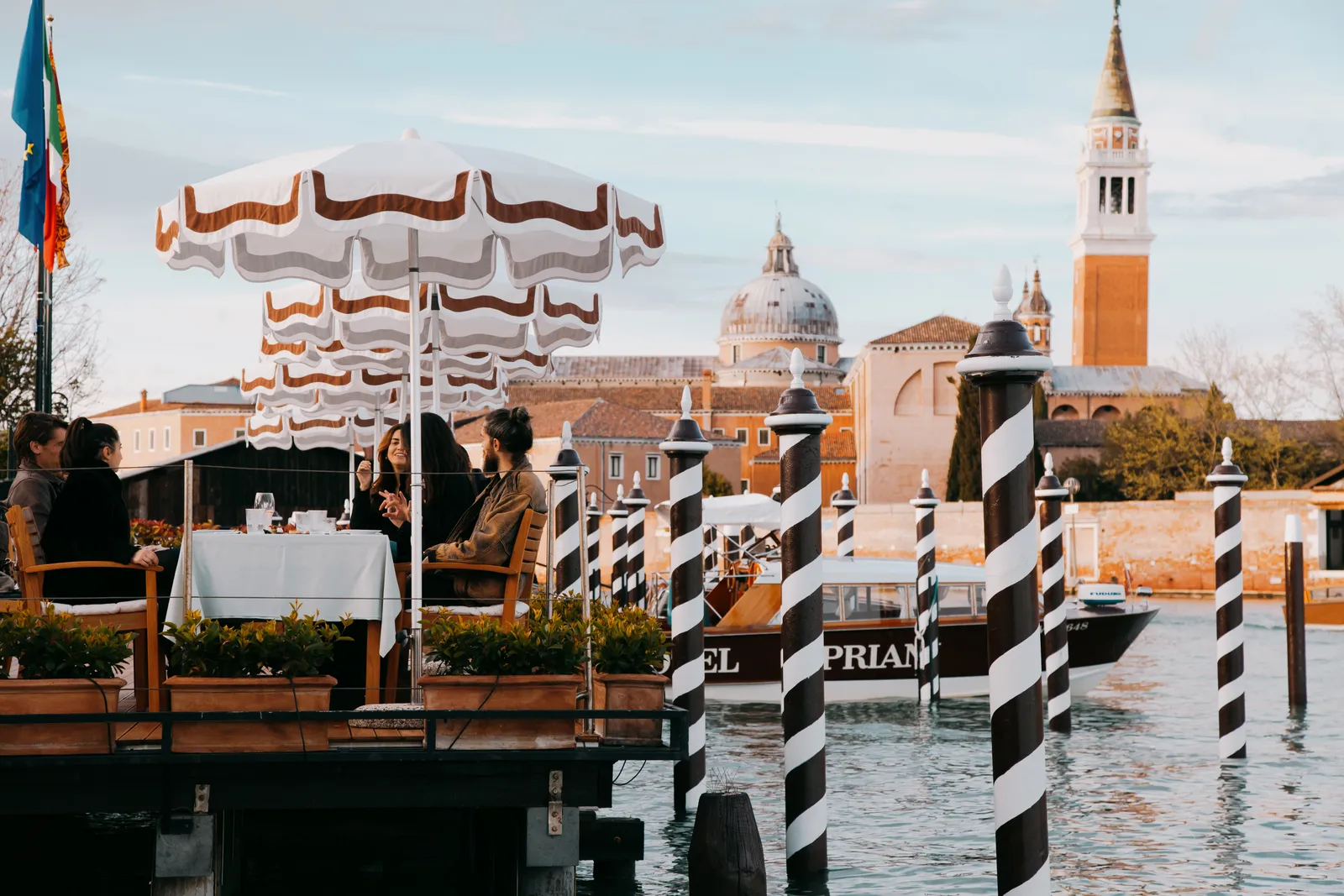 A table over the water under scalloped parasols, among the jetty’s striped poles, with San Giorgio Maggiore’s bell tower behind.