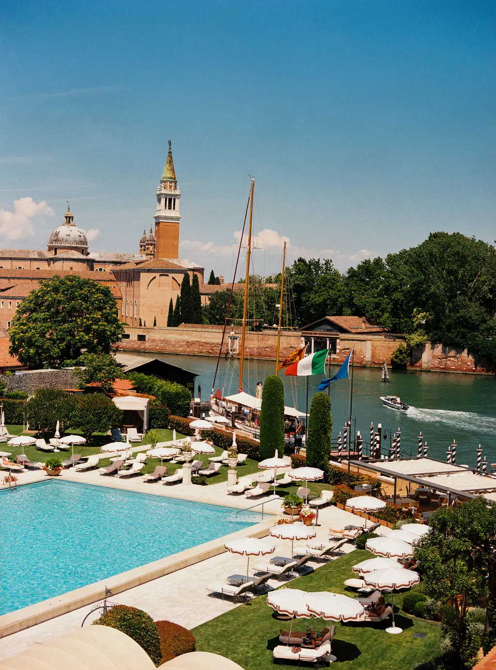 The pool among sunbeds and white parasols, with the bell tower and dome of San Giorgio Maggiore beyond the trees.
