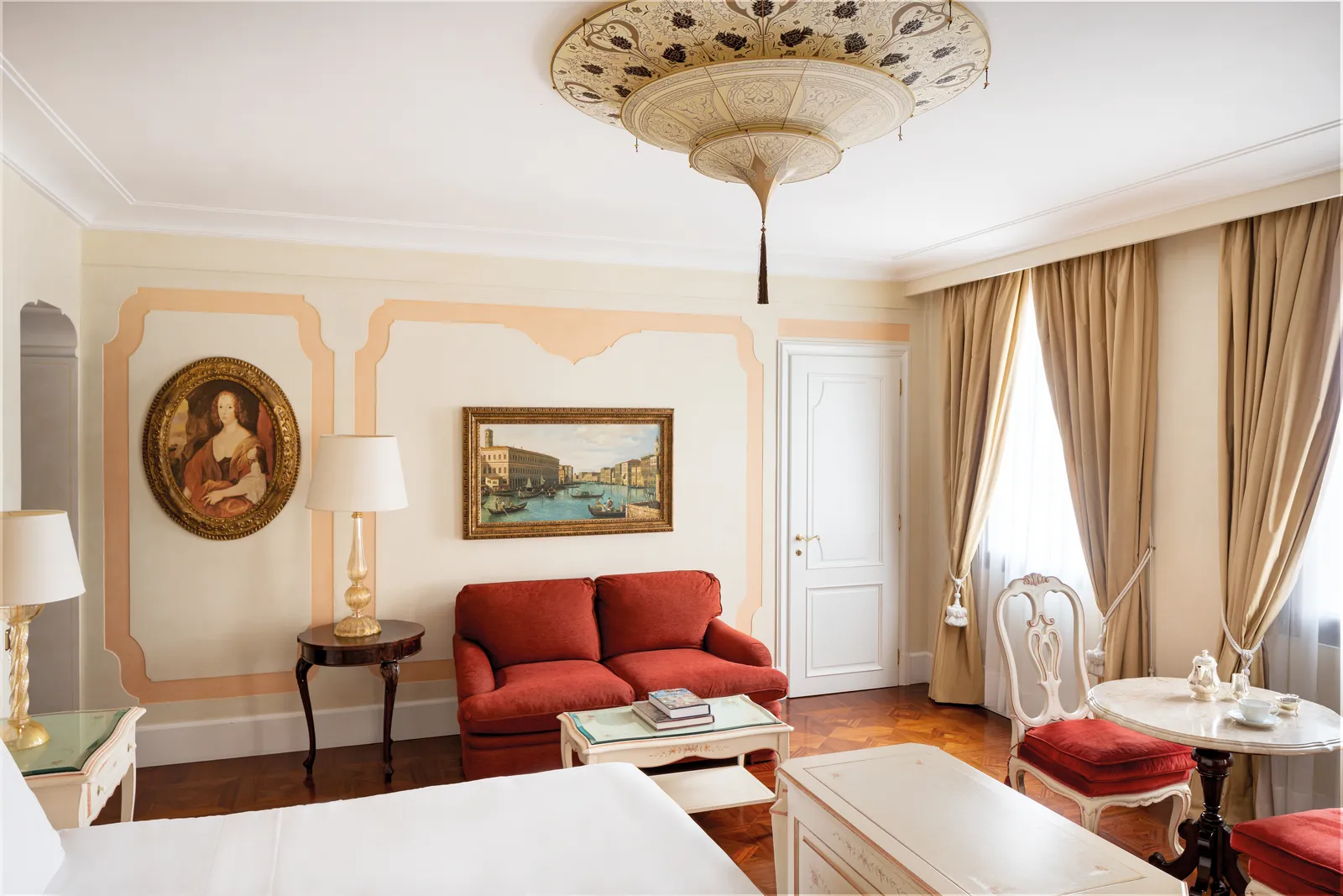 A junior suite with a red sofa, an oval portrait on the wall, a Fortuny lamp and sand-coloured curtains.