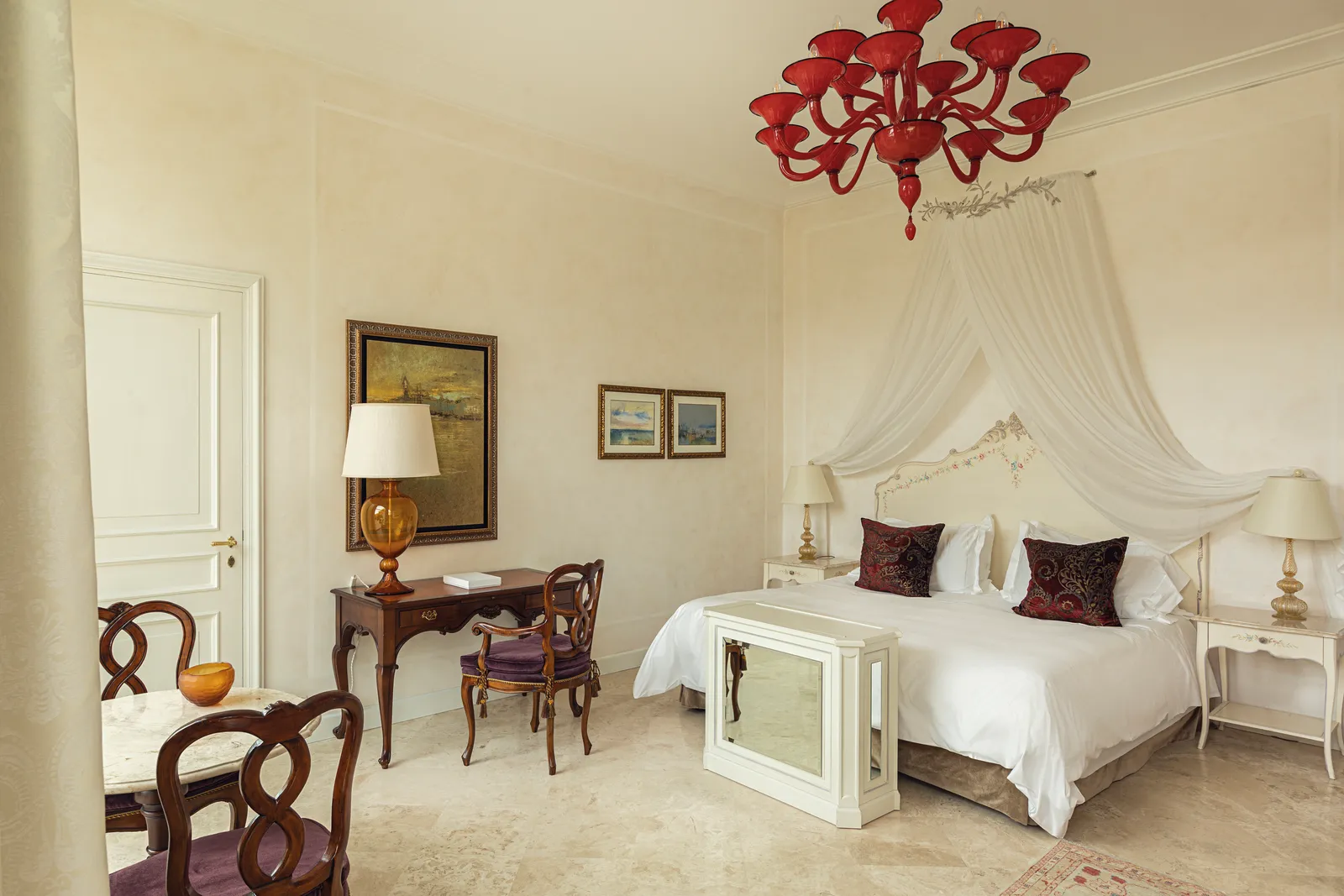 A junior suite with a tulle-canopied bed under a red Murano chandelier, a writing desk and dark chairs.