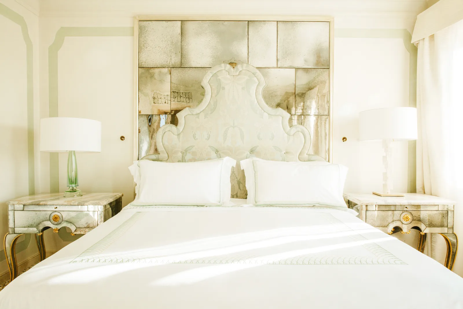 A white bed with a shaped headboard against a wall of antiqued mirrors, between 2 mirrored bedside tables.