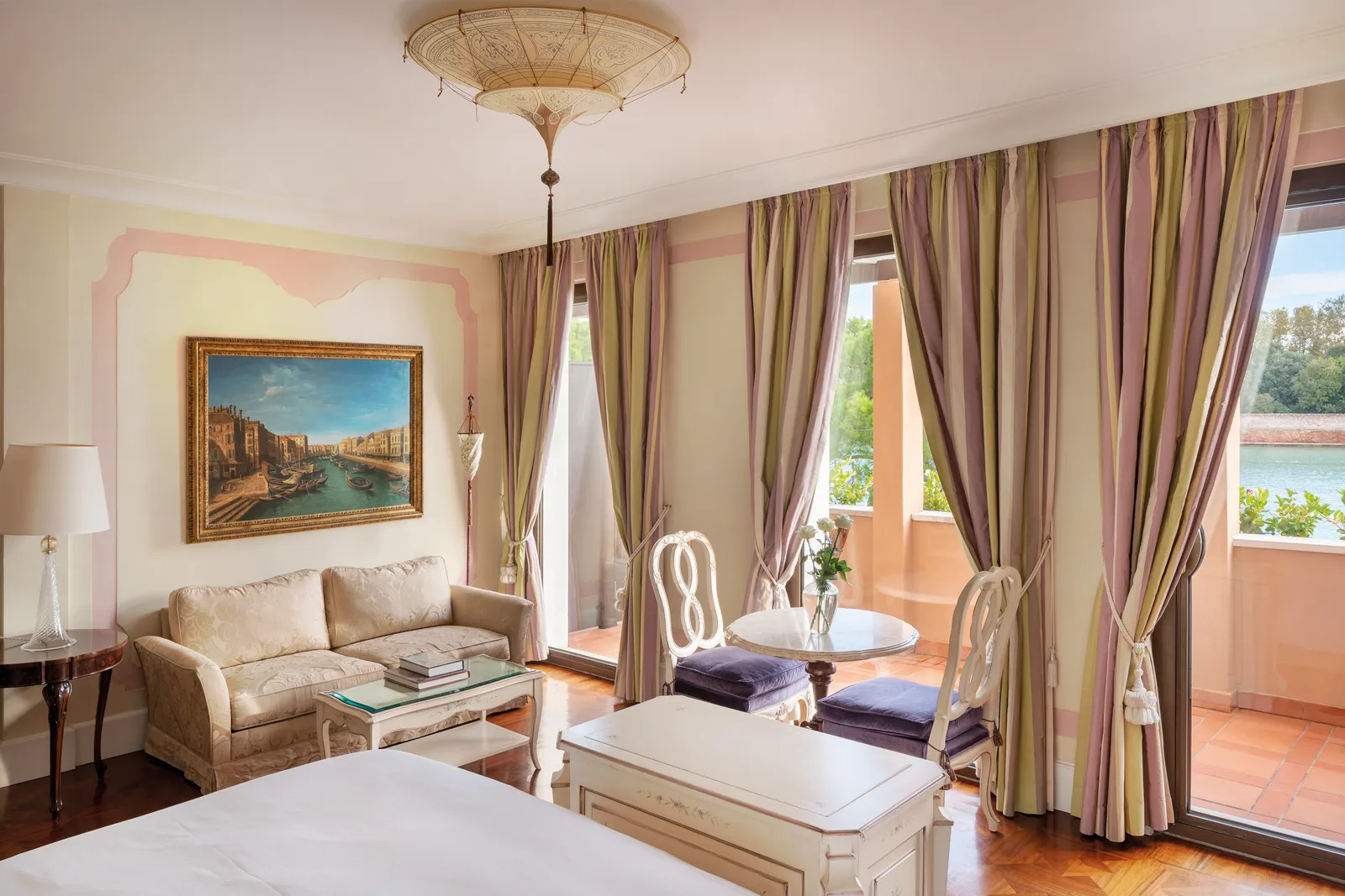 A room with a parquet floor, a small sofa under a view of Venice and striped curtains at the French windows onto the balcony and the lagoon.