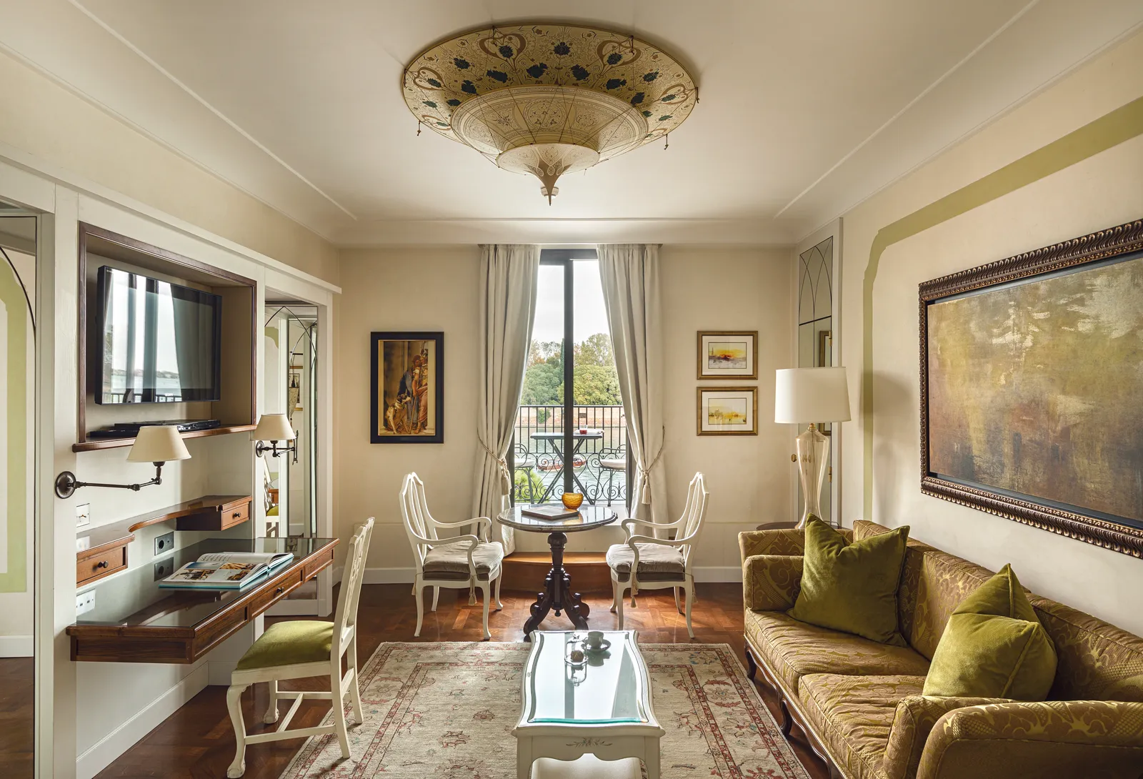 A suite’s living room with a green velvet sofa, a desk, a Fortuny lamp and the window onto the lagoon.