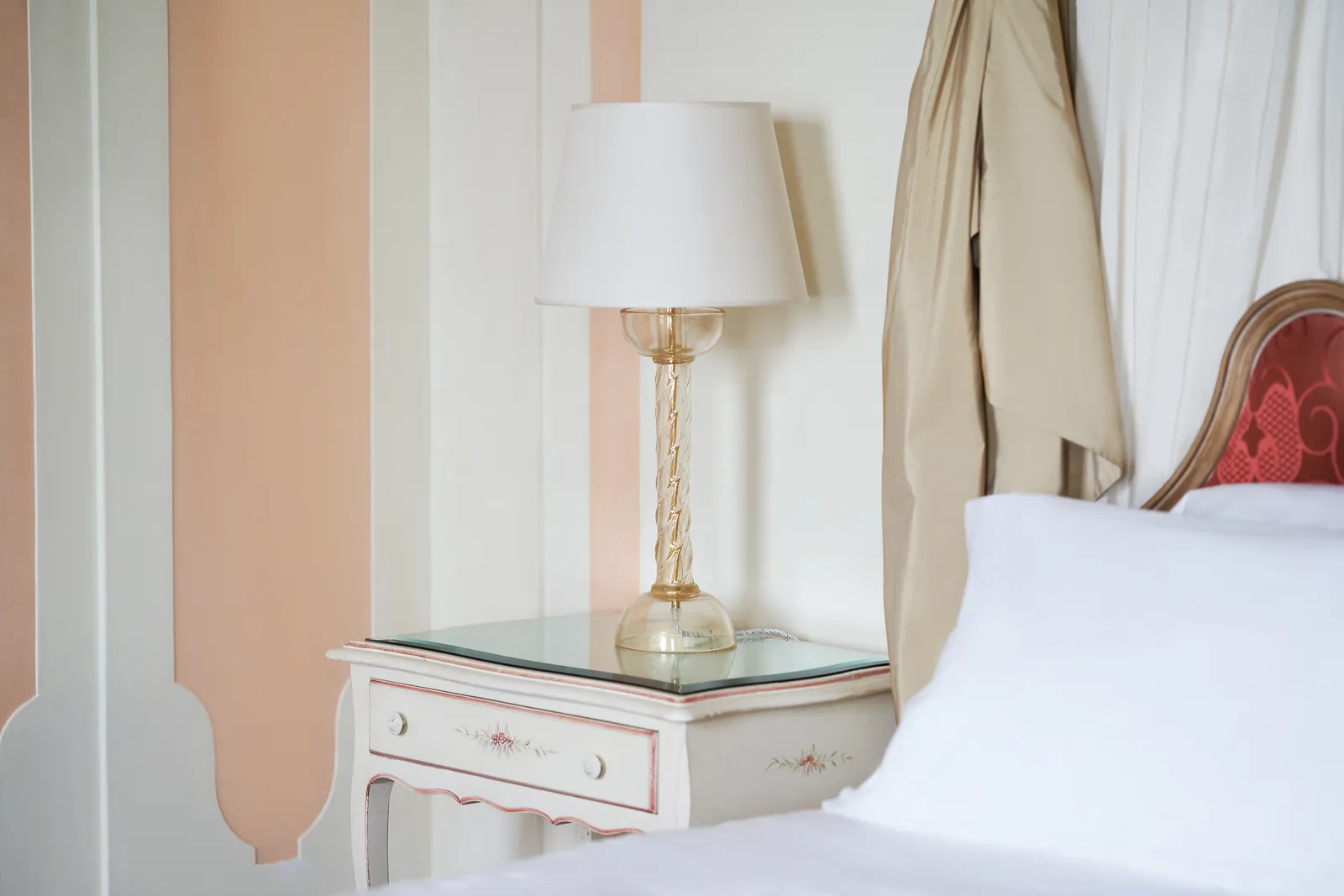 A painted bedside table with a Murano glass lamp, beside a bed with a red headboard.