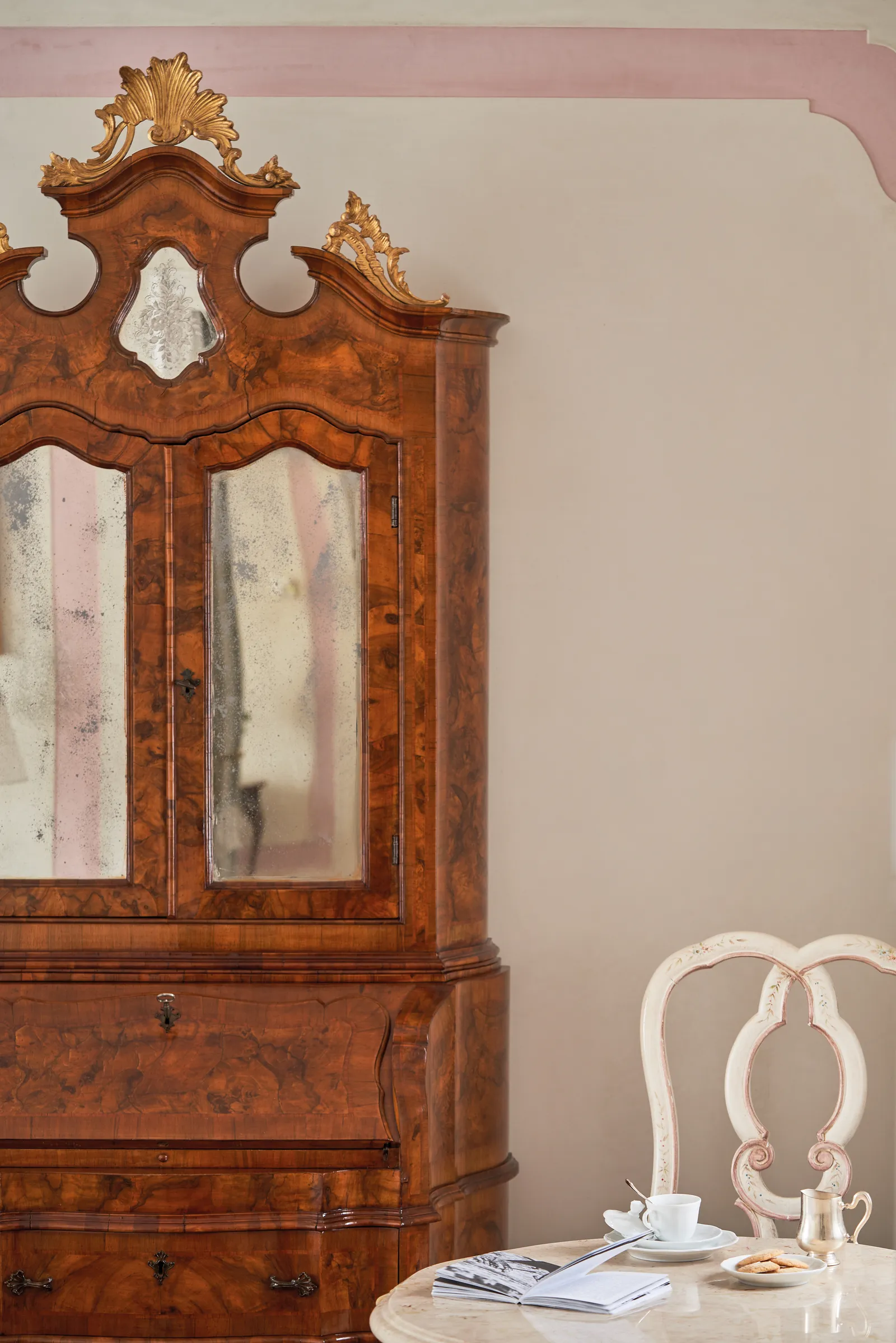 A Venetian walnut bureau cabinet with mirrored doors and, beside it, a white chair with a tea set.