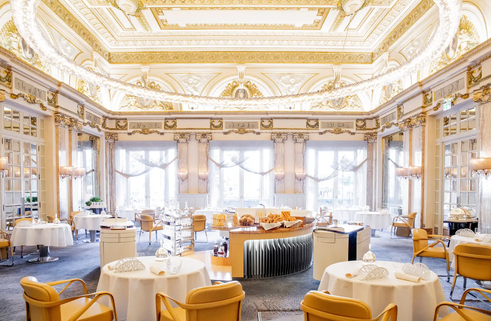 Le Louis XV’s dining room laid for service, with garland chandeliers, the gilded plaster ceiling and the French windows.