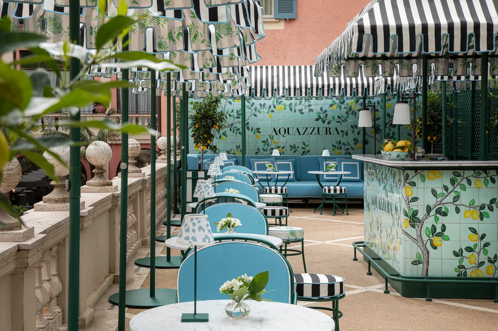 The Aquazzura Bar: awnings in black-and-white stripes and a botanical print, a counter and wall faced with tiles painted with lemon branches and the Aquazzura name, pale blue chairs and banquettes, small tables with lamps and a citrus tree in fruit