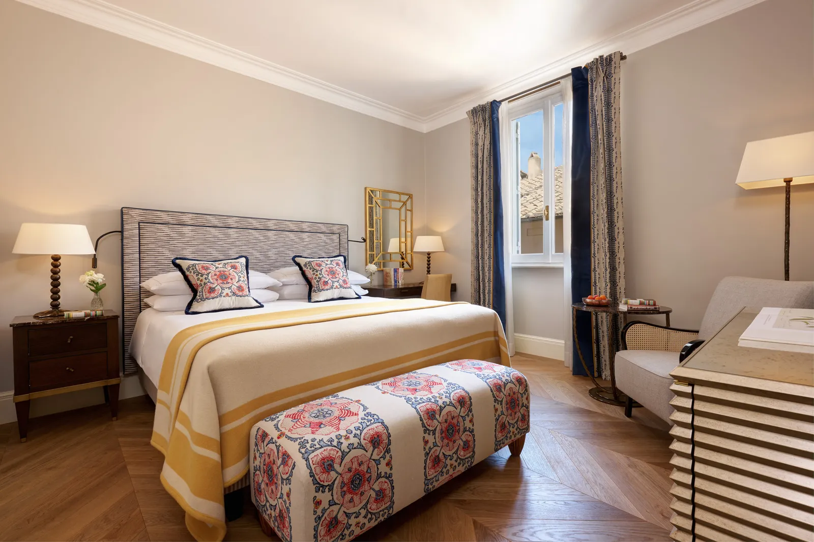 A Superior Room: a double bed with a yellow-striped throw and embroidered cushions, a floral bench at its foot, herringbone parquet and a window onto the rooftops