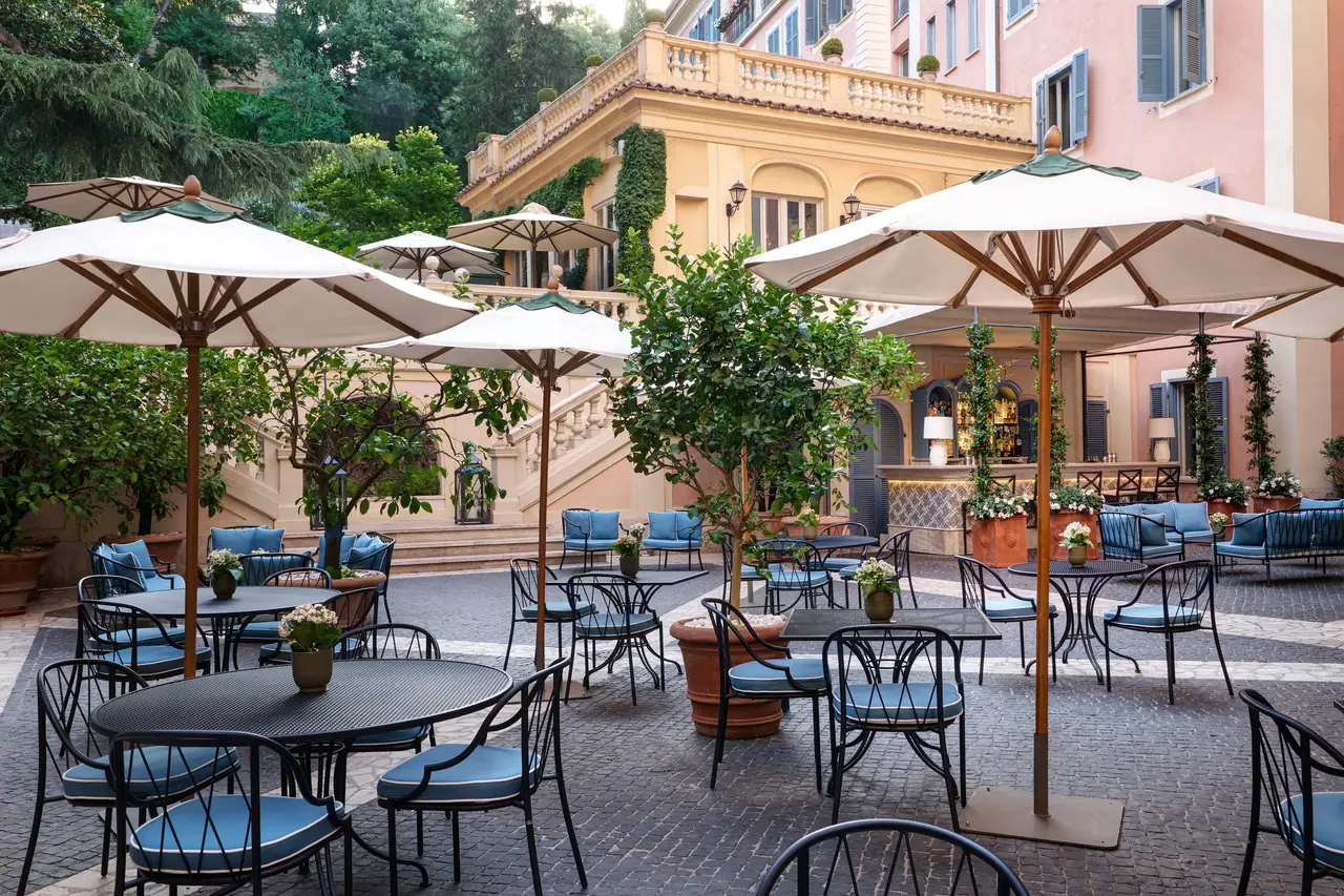 The Stravinskij Bar courtyard by day: wrought-iron tables and chairs with pale blue cushions, cream parasols and small trees in terracotta pots on the cobbles; the counter under its awning at the back, a balustraded staircase and an ivy-covered wall on the left