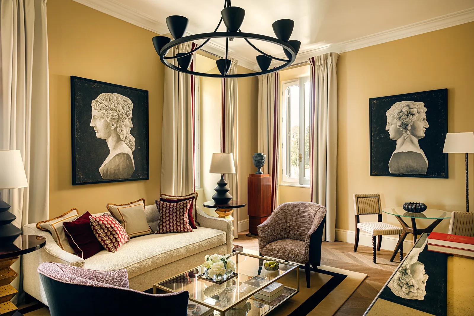 The sitting room of a Deluxe Suite: ochre walls with 2 large prints of classical heads, a black chandelier, a pale sofa with burgundy cushions, a glass coffee table with white flowers and windows with red-striped curtains