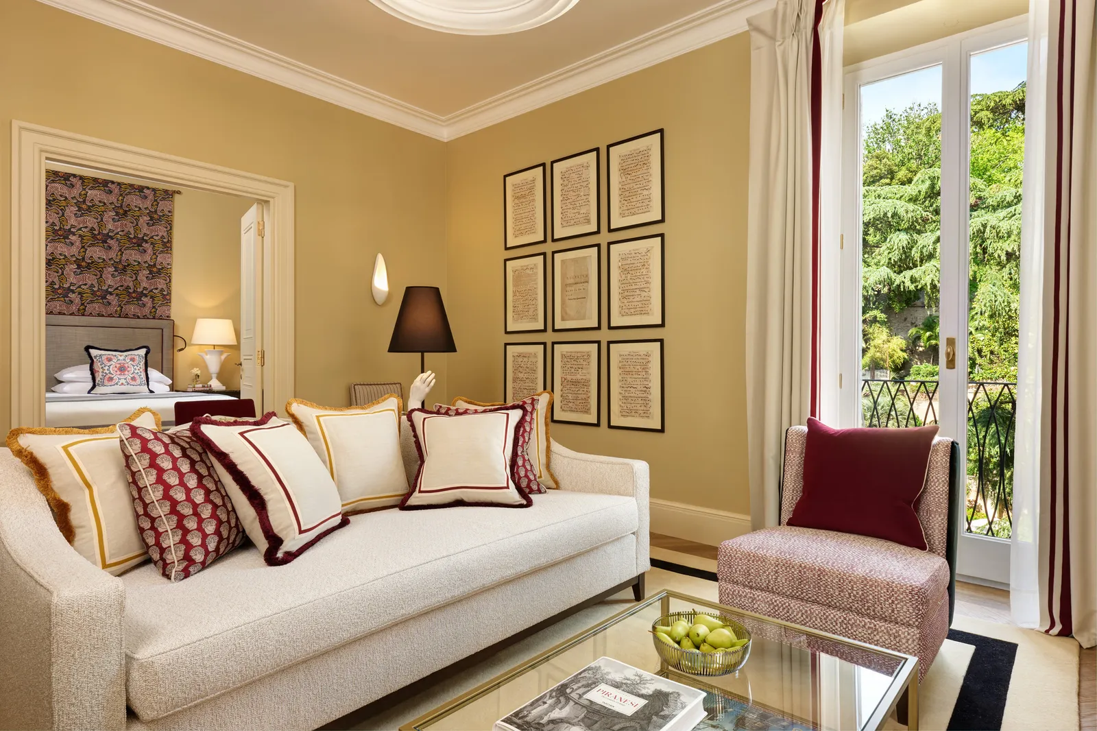 The sitting room of a Deluxe Suite Garden View: a pale sofa with red and white cushions, 9 framed manuscript pages on the wall, the French window open onto a small balcony and the garden’s trees, and the bedroom through the door