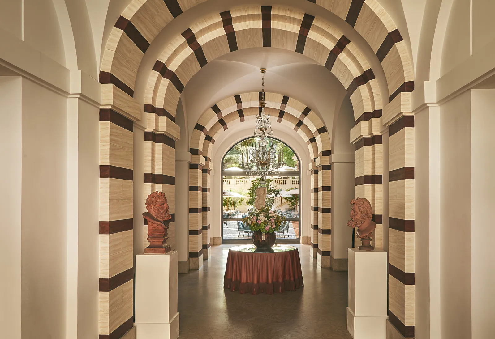The lobby gallery: arches striped in cream and brown, 2 terracotta busts on white plinths, a round table with a large vase of flowers and, at the end, the arched doorway open onto the courtyard