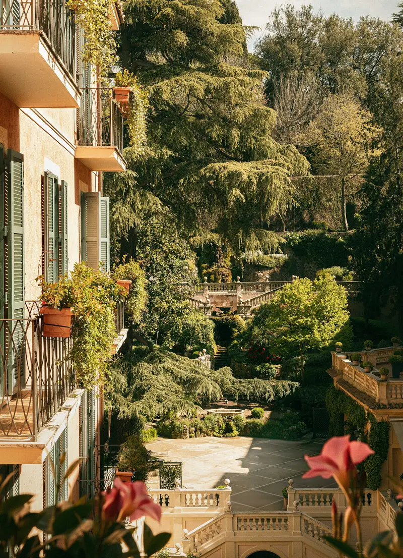 The Secret Garden from one of the hotel’s balconies: the façade with green shutters and flowering pots on the left, pink flowers in the foreground, a paved terrace below and balustraded terraces rising among tall trees