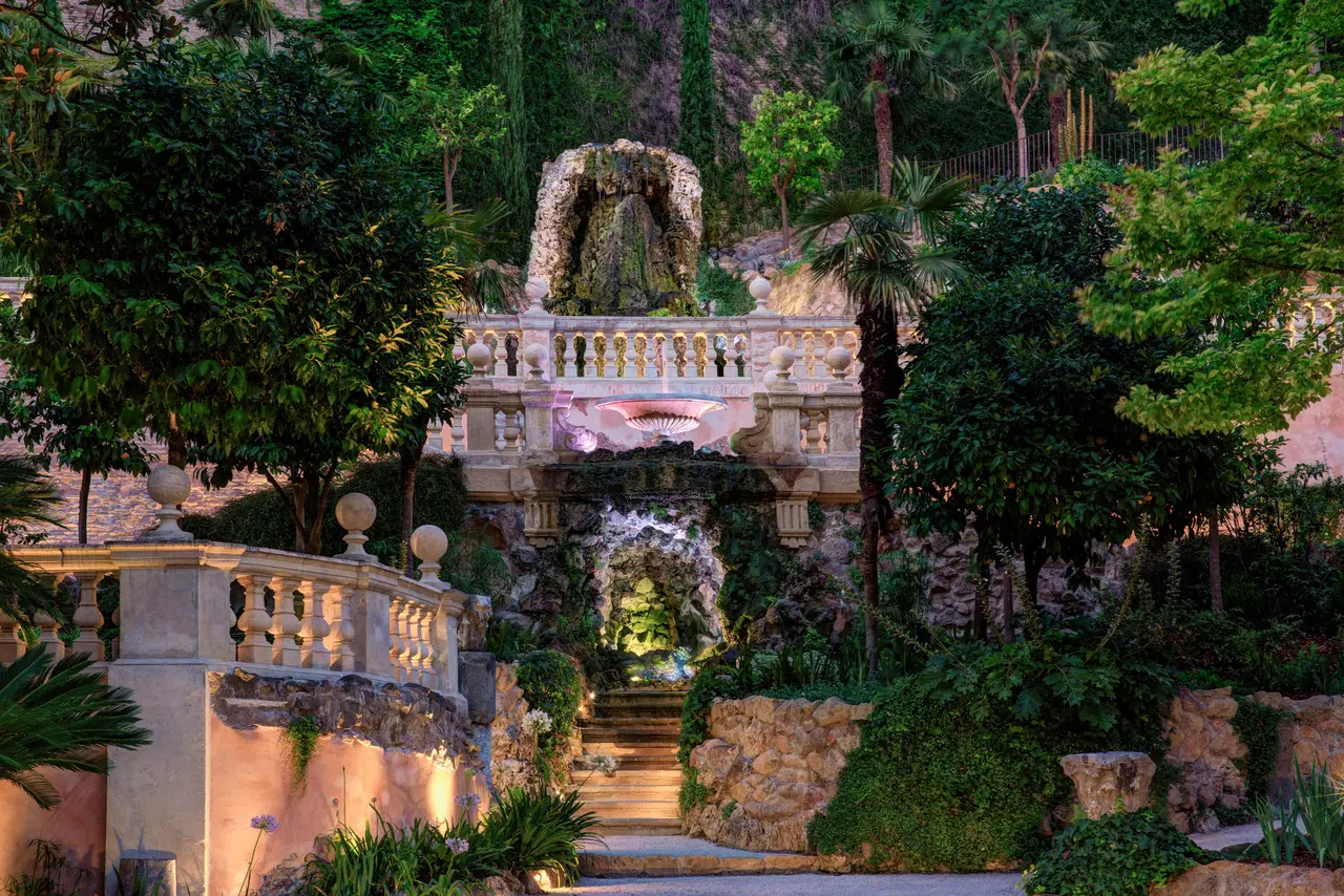 The Secret Garden in the evening: a balustraded terrace topped by a rustic stone arch, with a lit grotto and a stairway below, among palms, trees and rocks